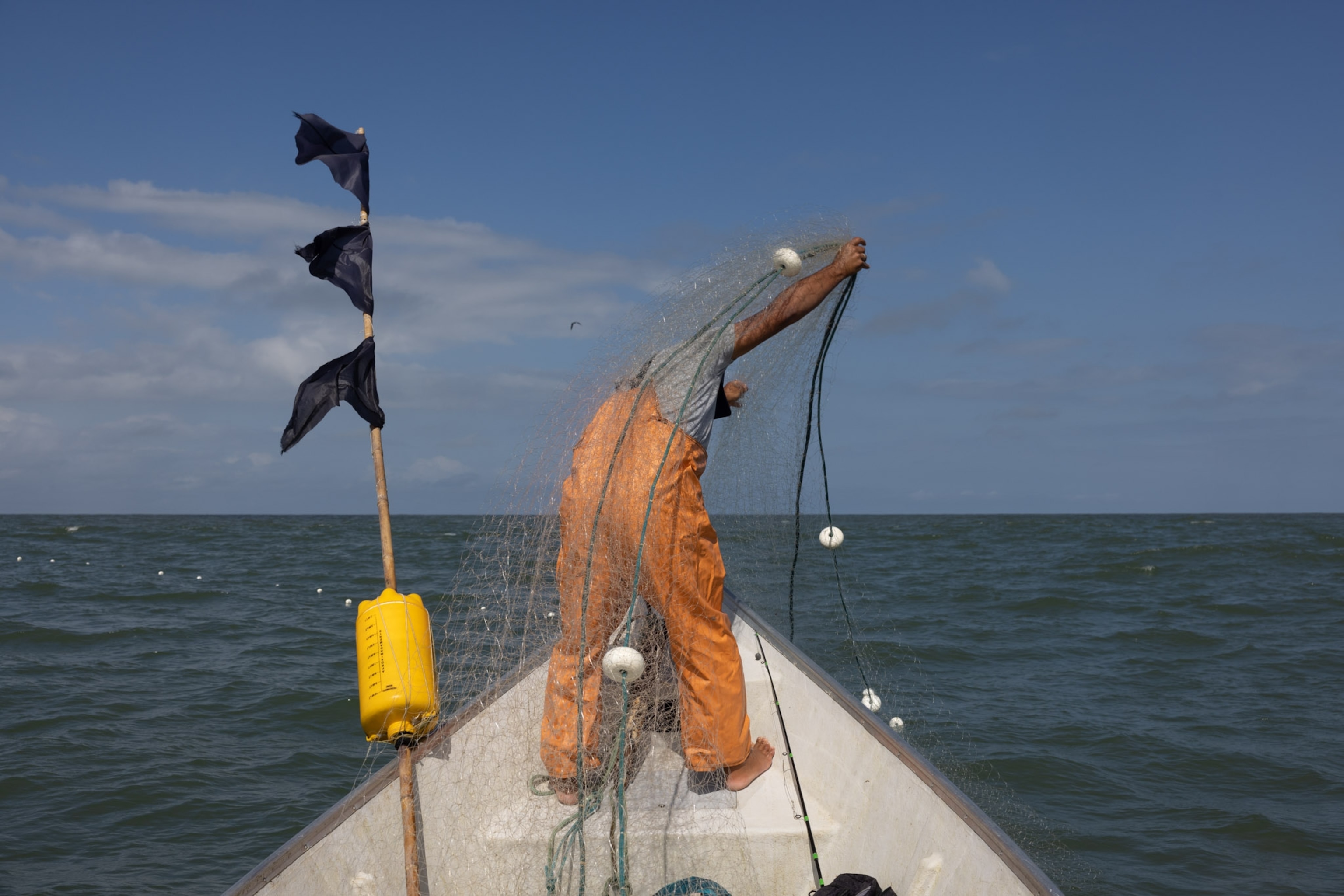 A fisherman in orange stands on the boat with net.