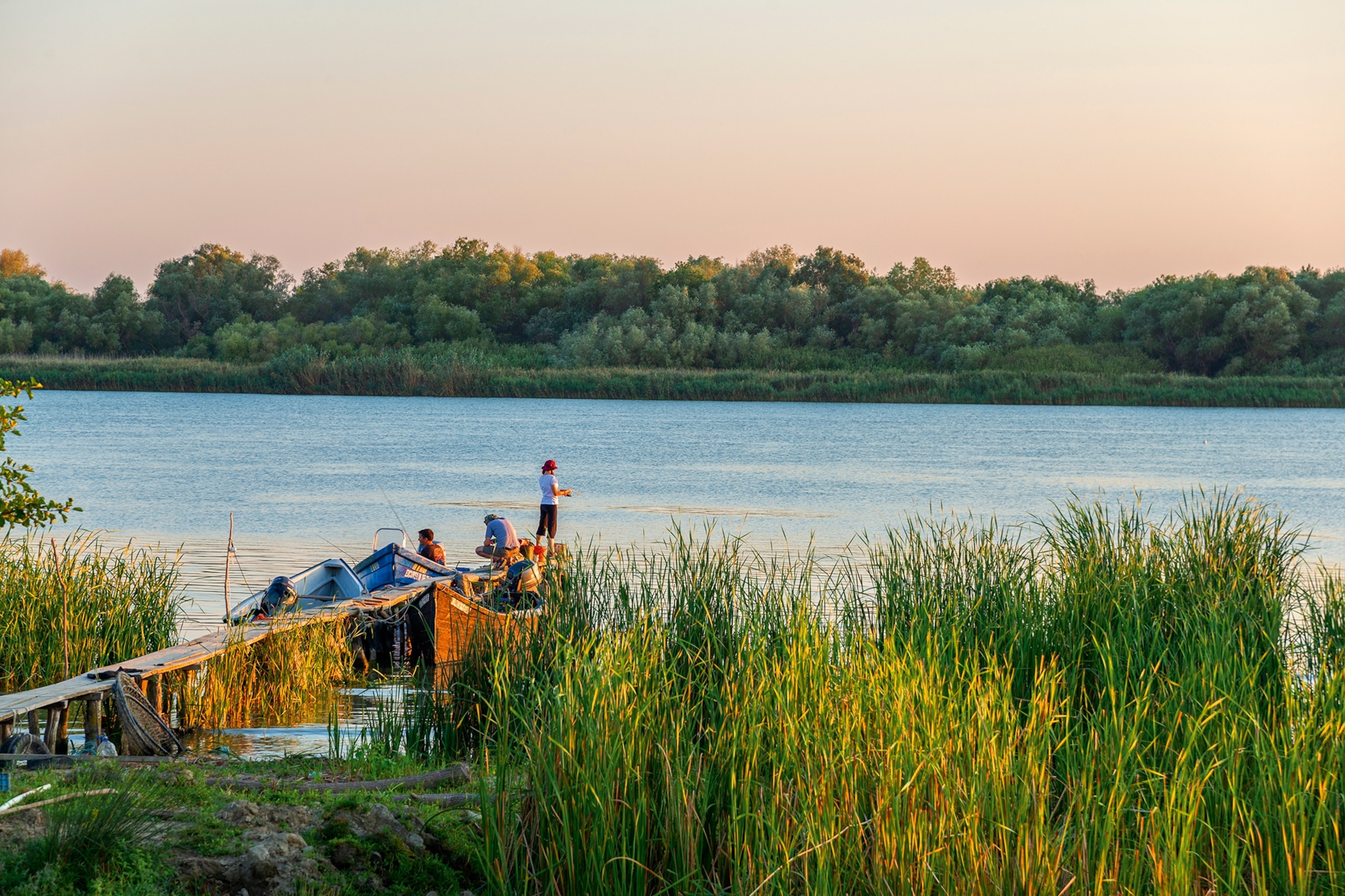 A small group of people fishing at the end of a pier at sunset surrounded by river boats.