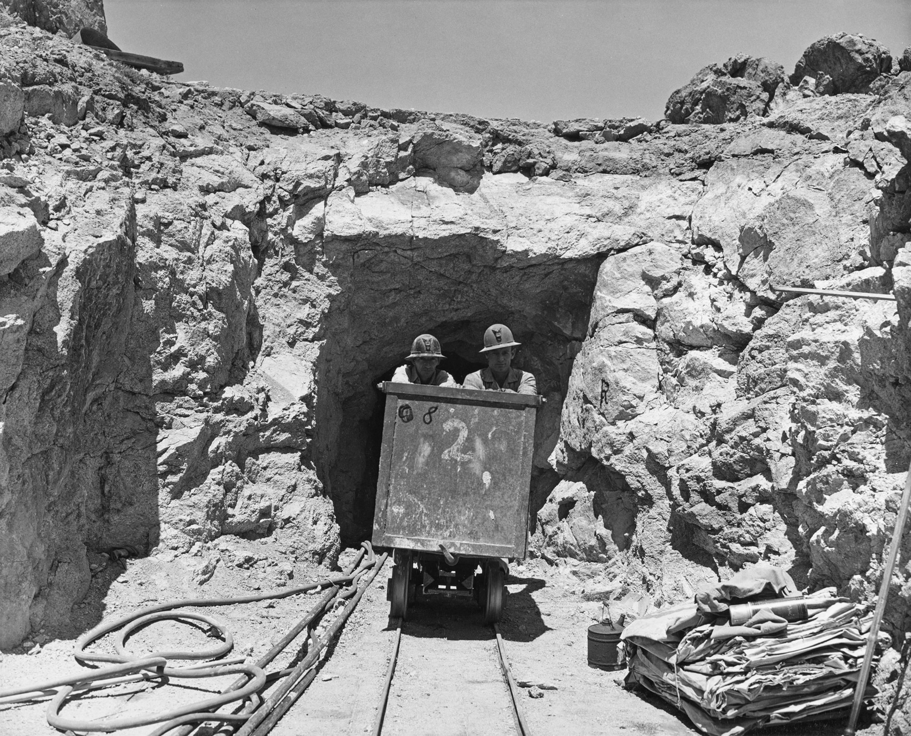Two miners, both wearing a hardhat, pushing an ora cart as they emerge from an uranium mine