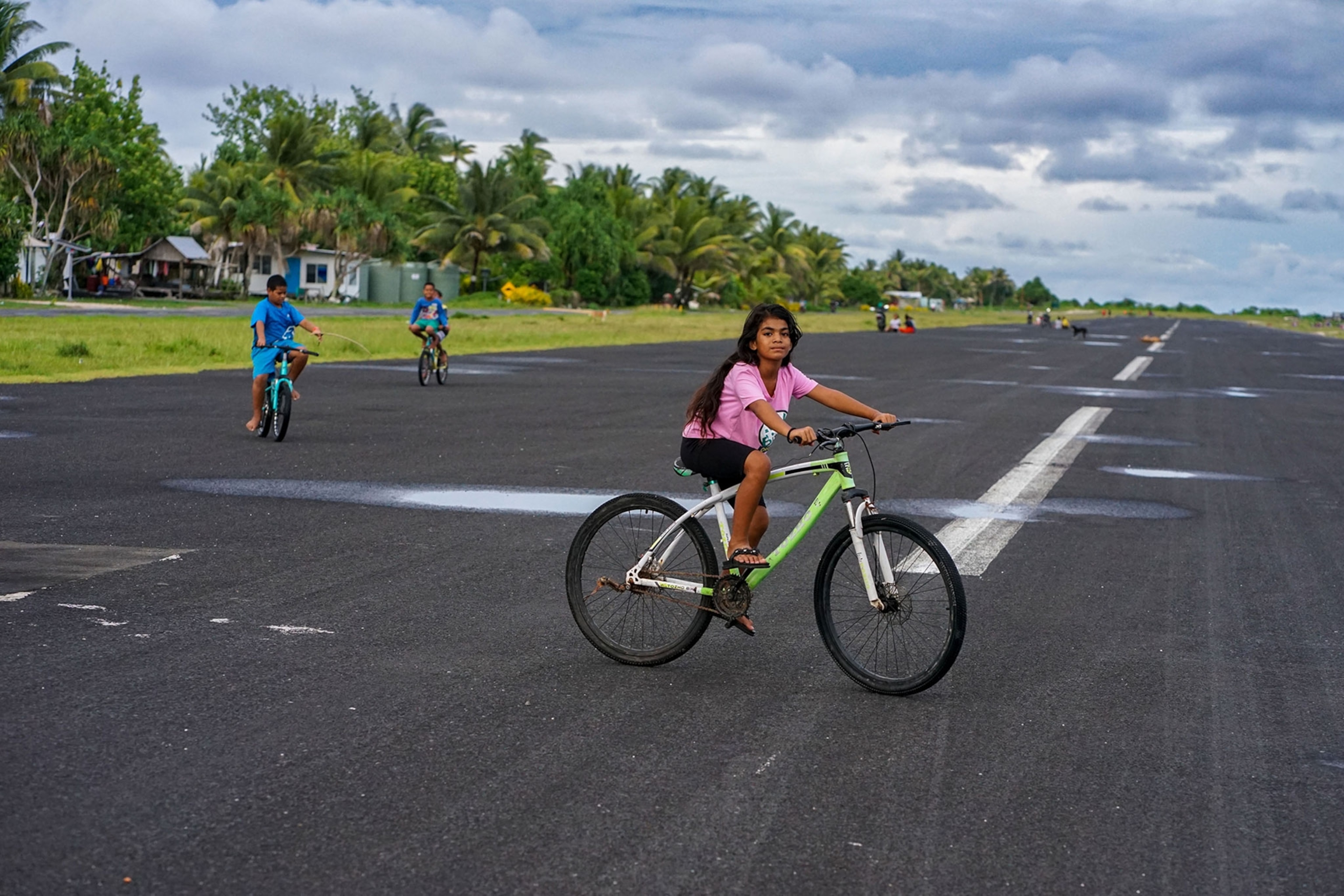 A young girl riding her bike on an airstrip on the island of Funafuti in Tuvalu stops to look at the camera. In the background of the image more children ride their bikes on the airstrip in front of a line of green trees and houses.