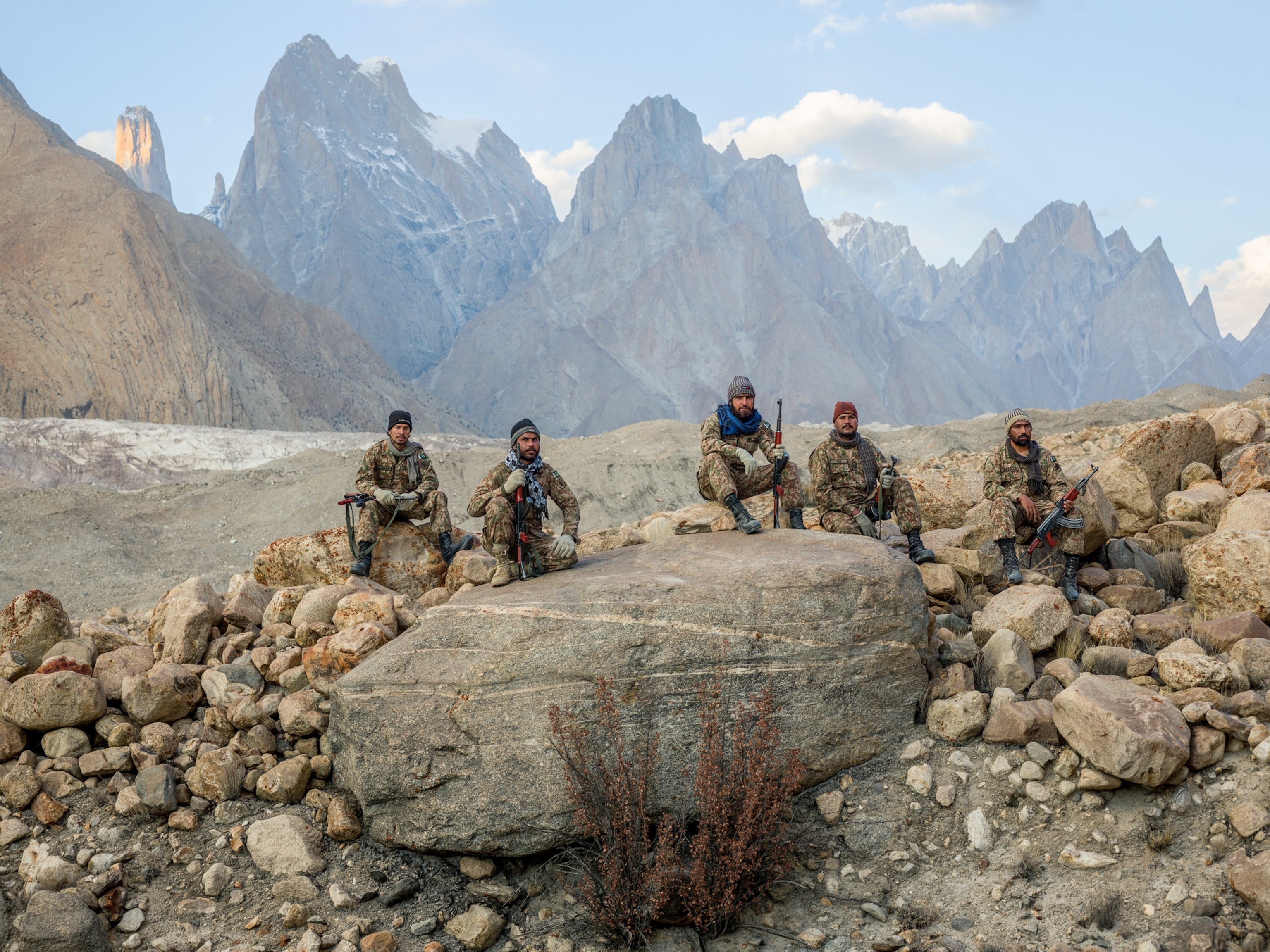 five soldiers sitting by large boulder with mountains on background.