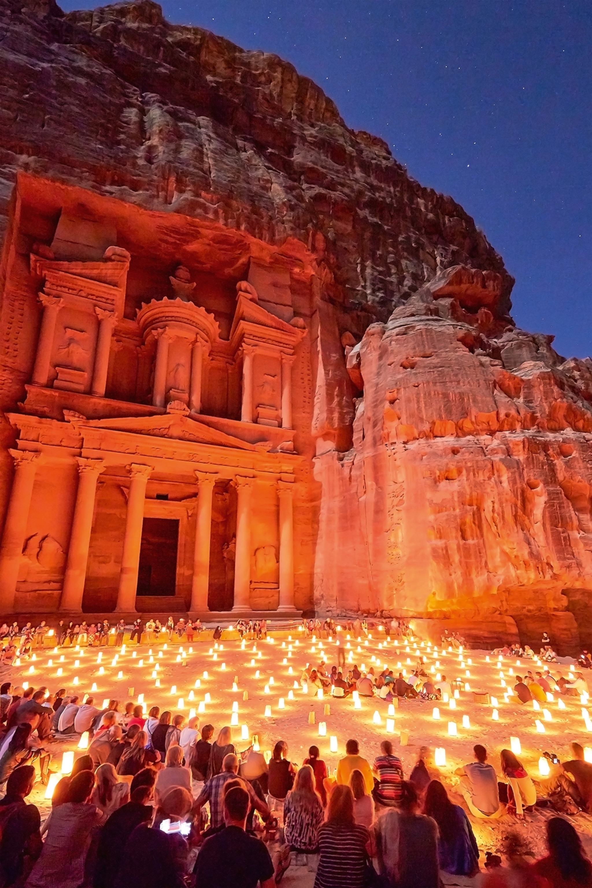 An ancient temple etched into a rock formation at night, with visitors sitting around countless little lamps in the sand in front.