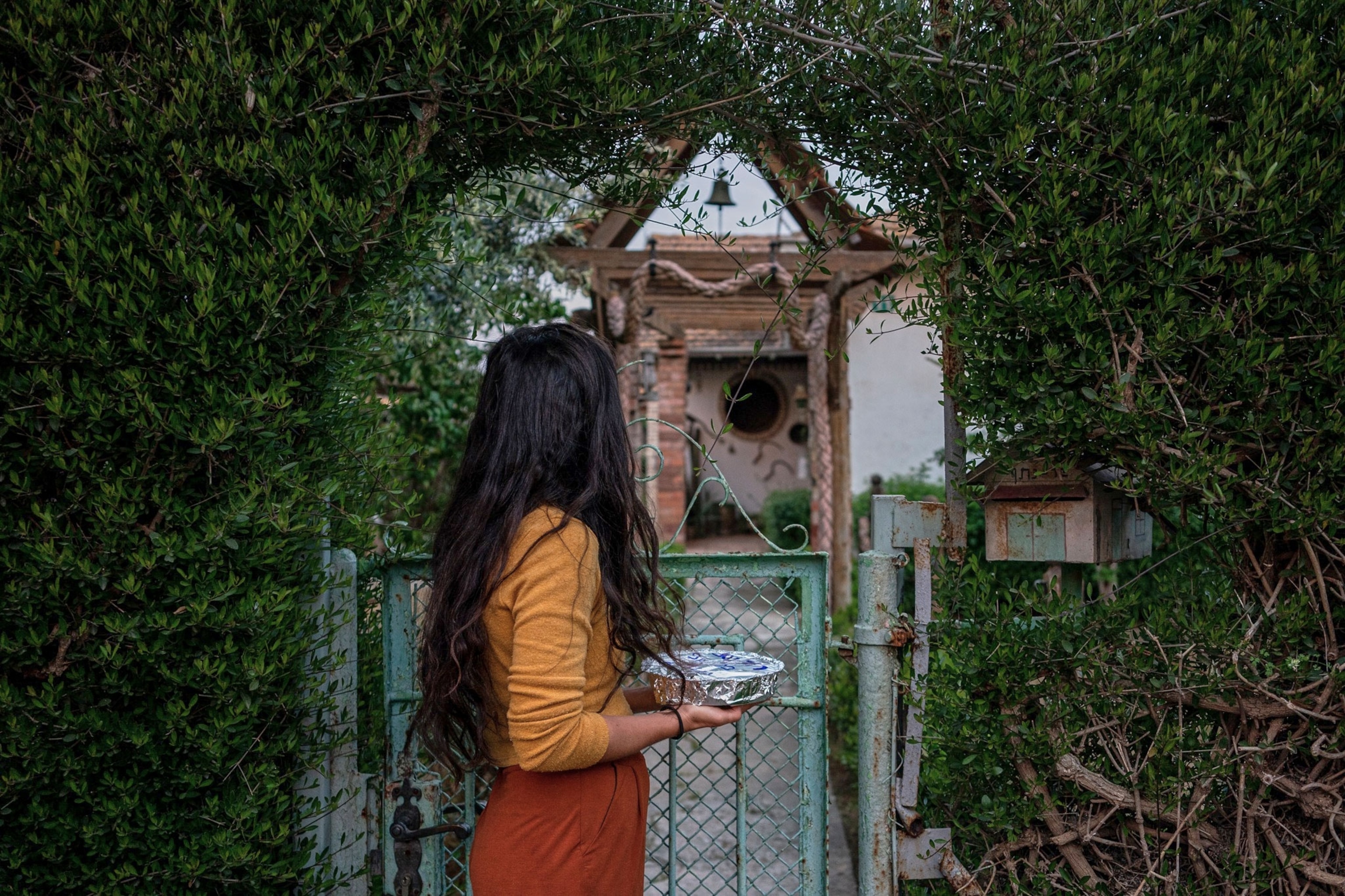 A woman stands in front of a houses gate that is surrounded by leaves. The woman stands facing away from the camera, looking towards the front door of the house while holding a foil container of food.