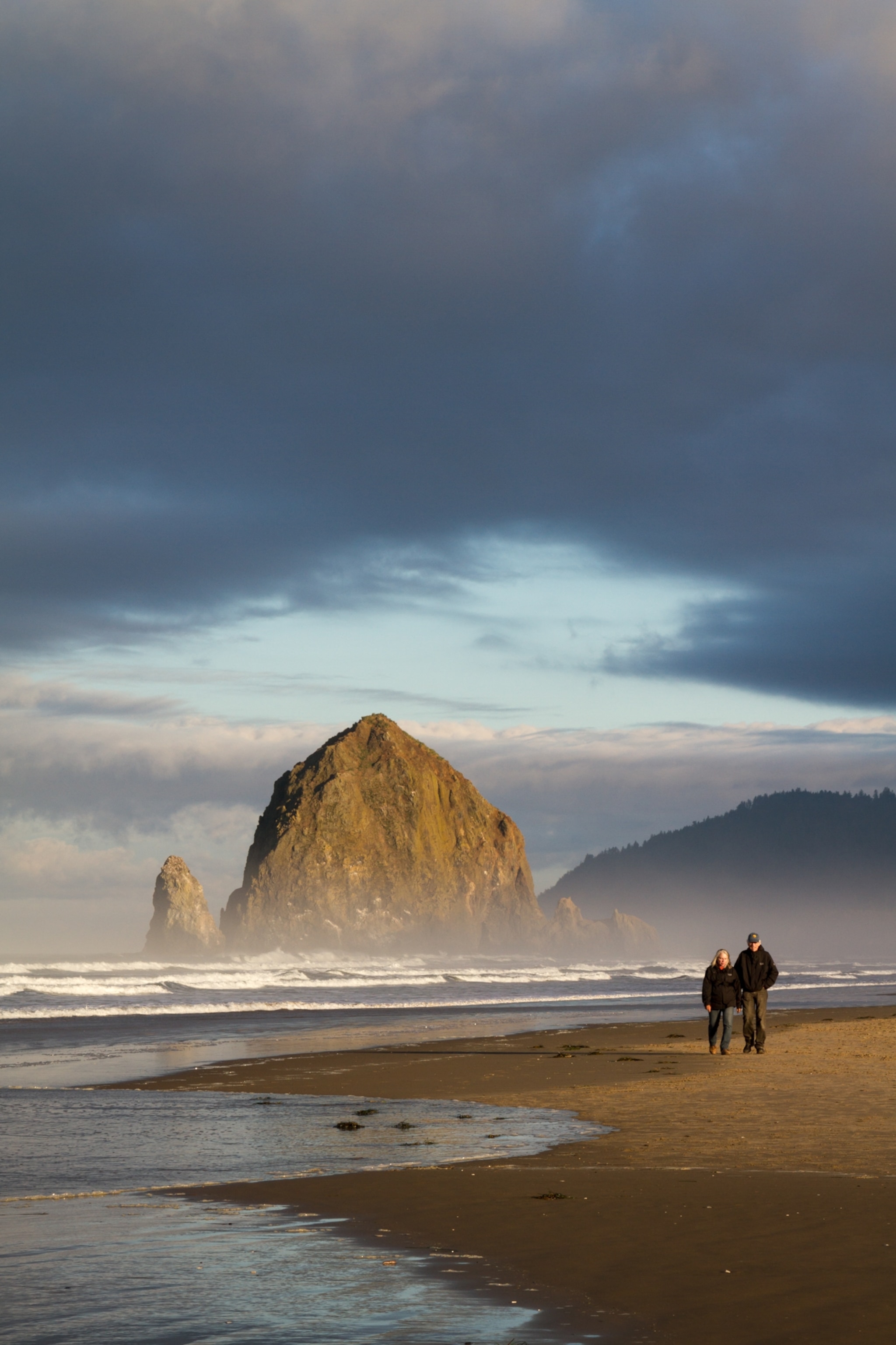 A couple walking along the beach with an enormous sea stack in the background.