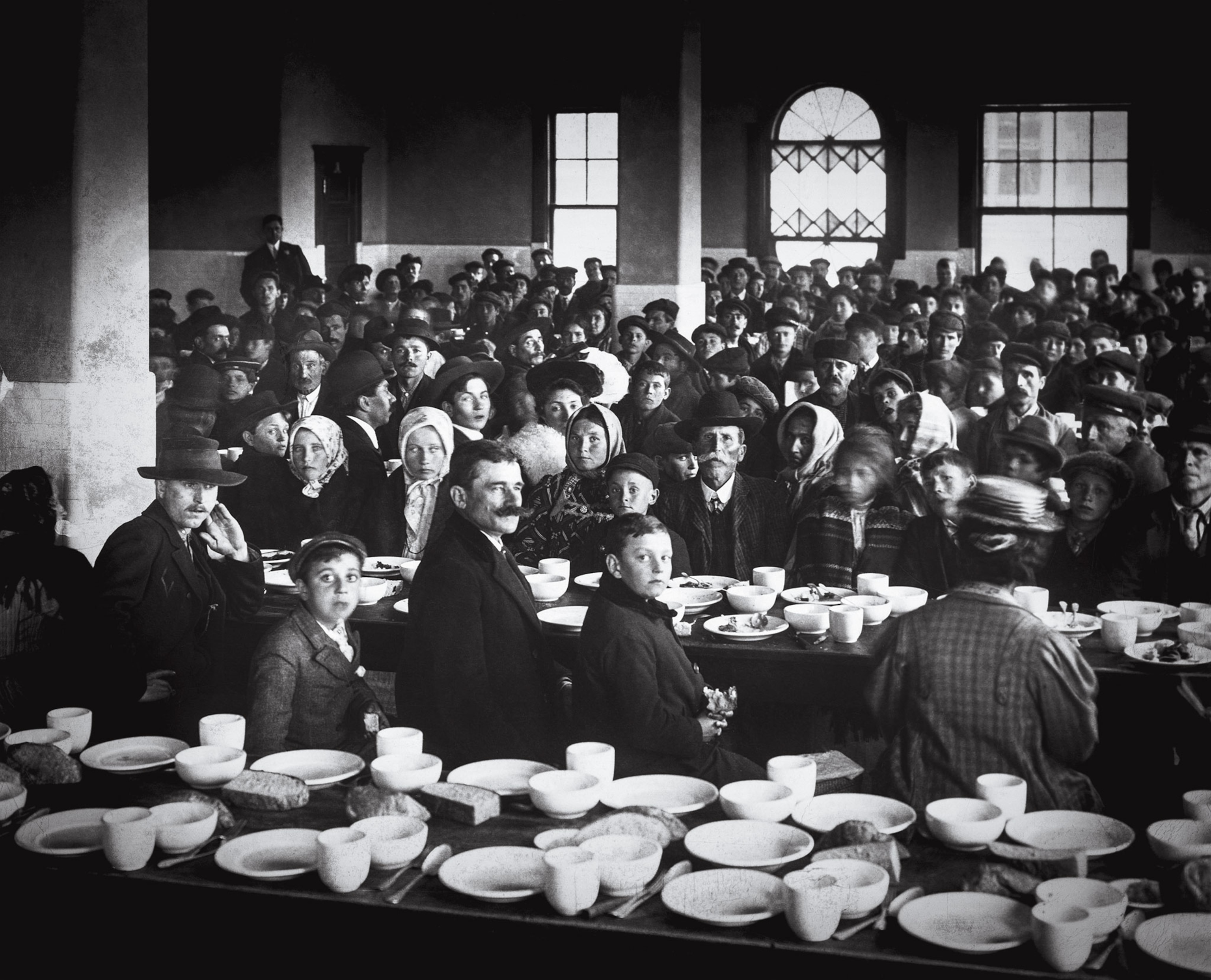 A black-and-white image of immigrants in the Ellis Island dining room, with the tables covered in white dishes.