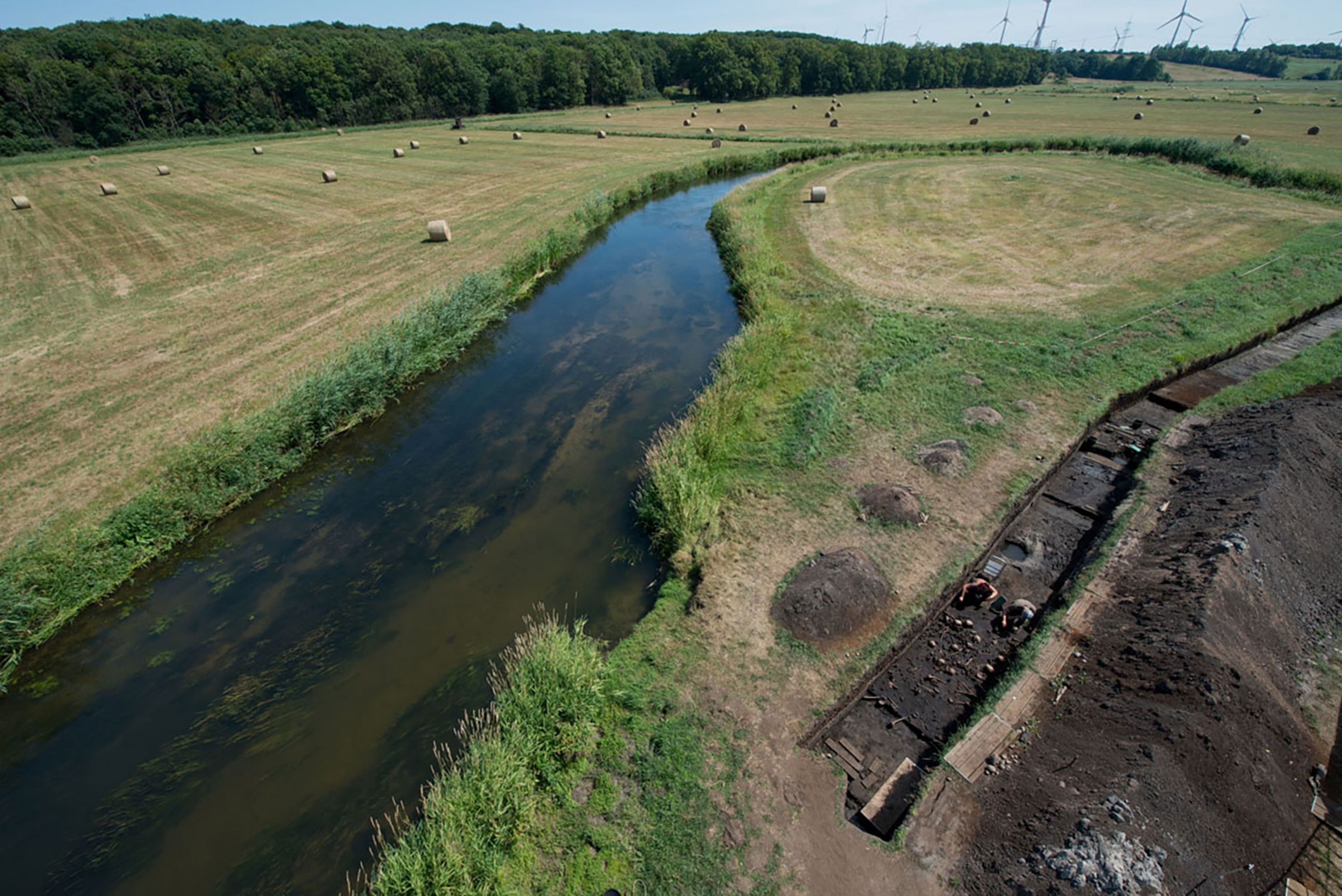 Aerial view of green field with river running through center of the frame. Excavation site running alongside the river to the right of the frame.