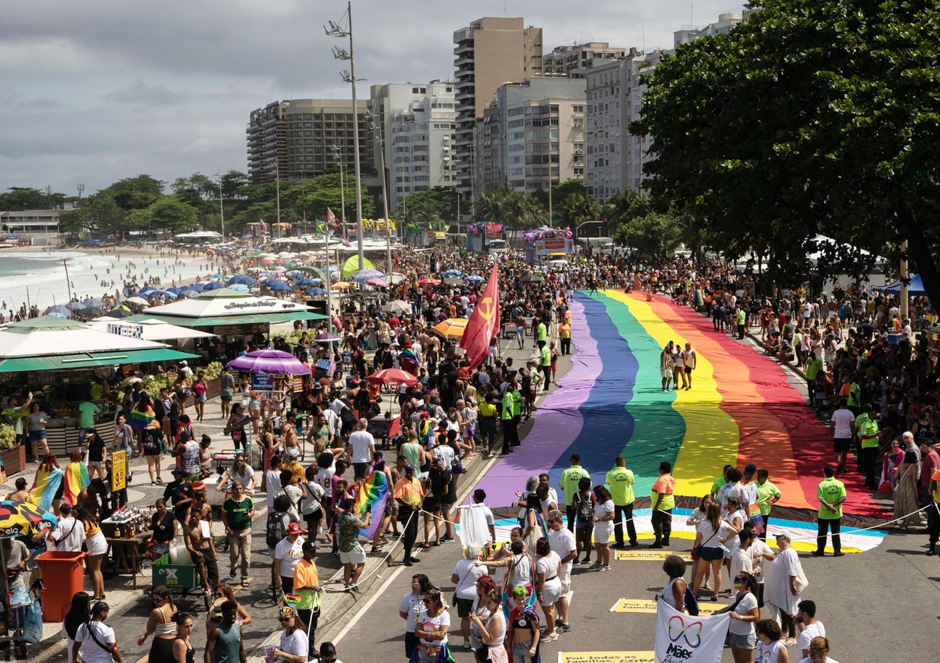 Revelers take part in the Pride Parade at Copacabana beach in Rio de Janeiro, a large rainbow flag blankets the street.