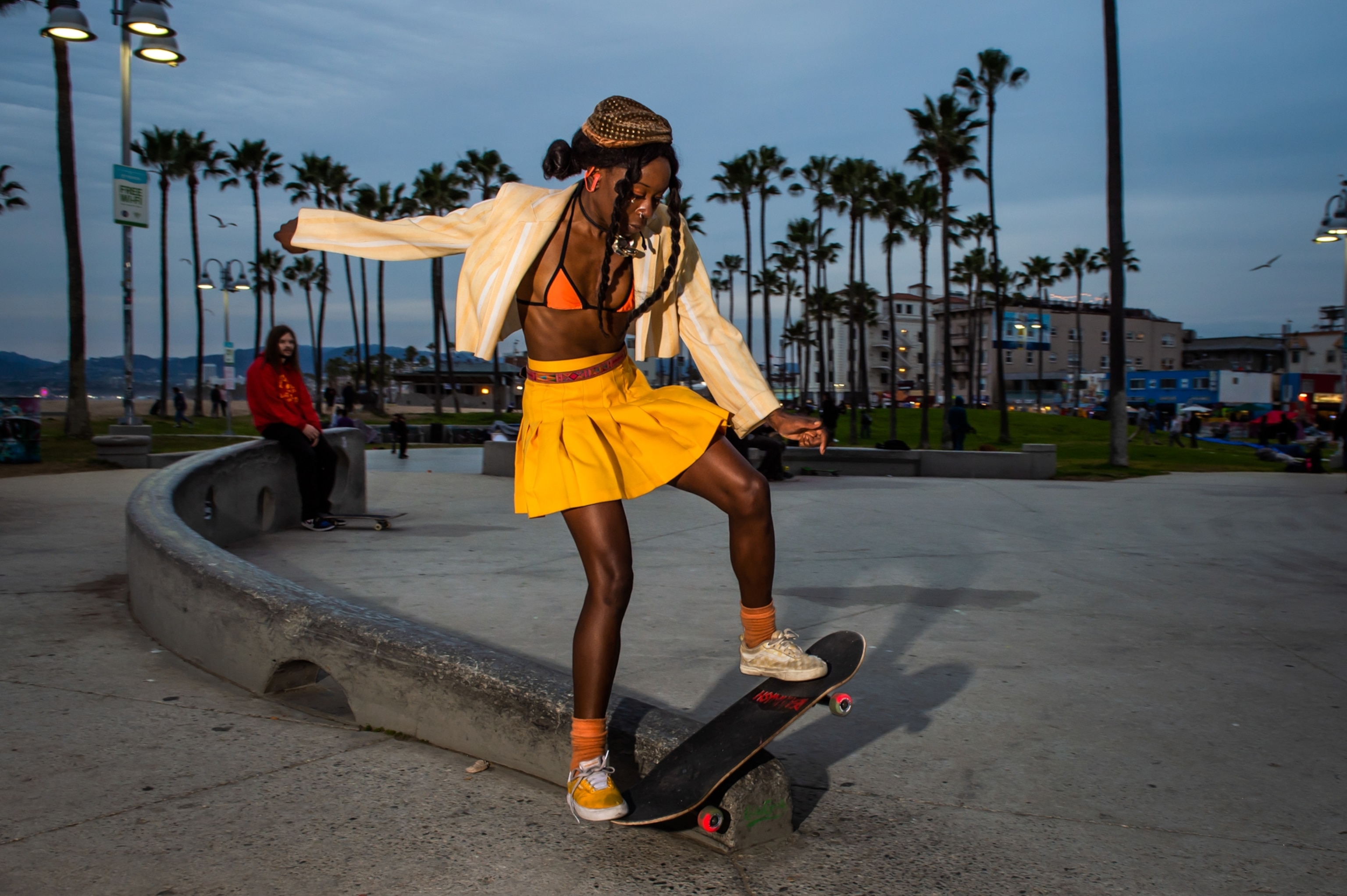 A young woman dressed in an unbuttoned yellow and white striped blazer, yellow pleated skirt, and yellow sneakers board-sliding down a waxed ledge.