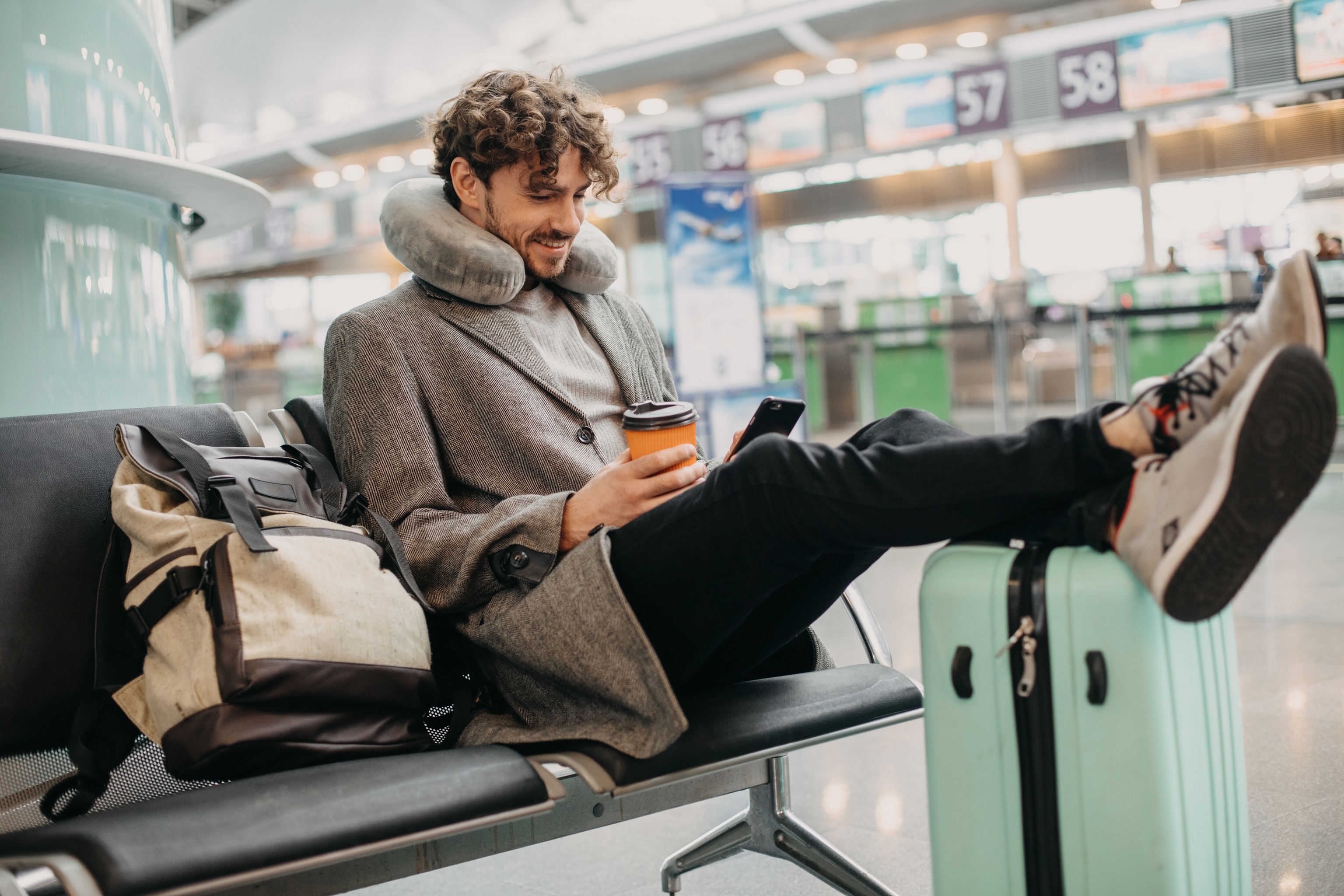 Man sitting in airport with feet up on suitcase and neck pillow around neck.