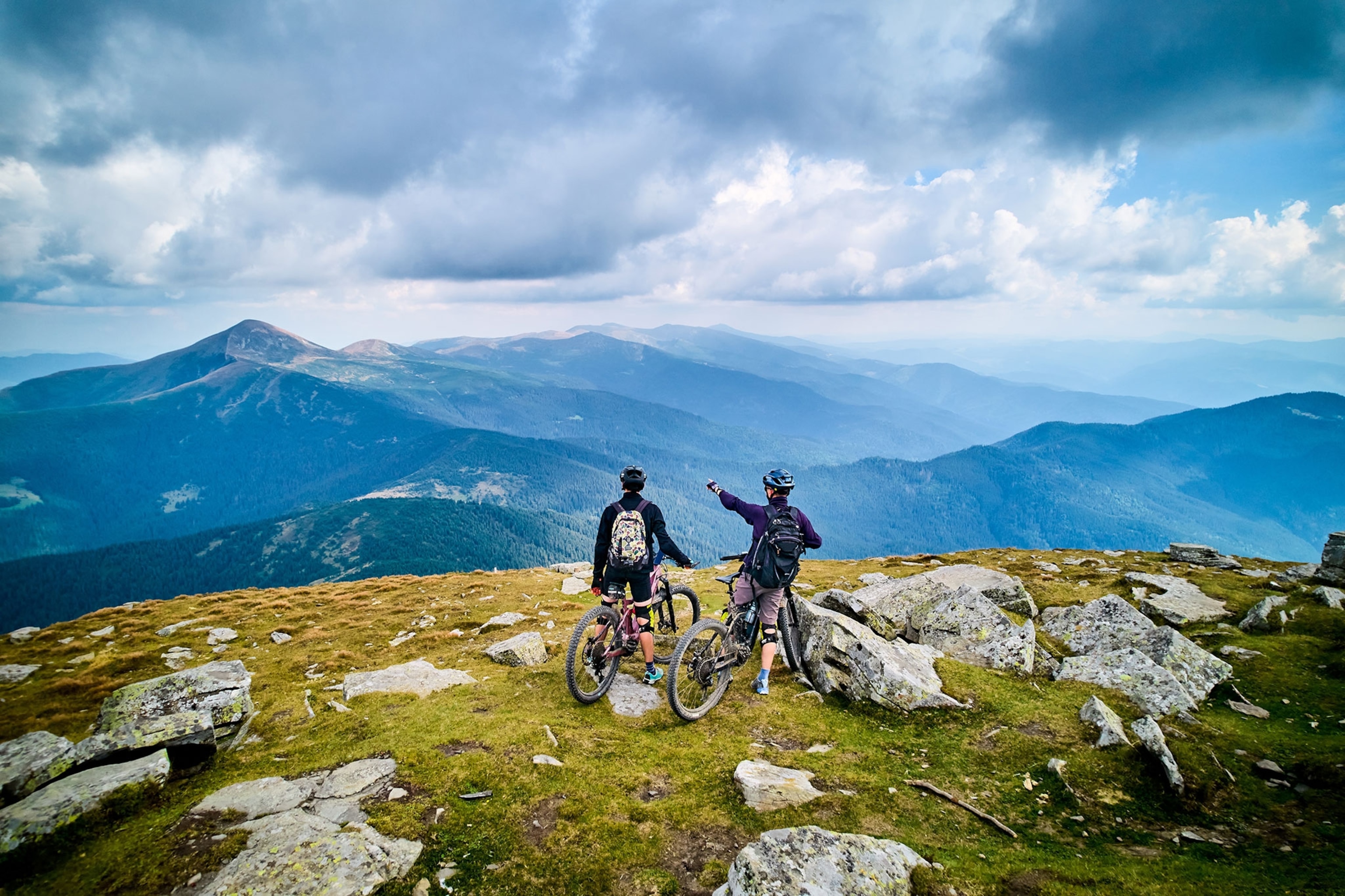Two people on e-bikes on a hilltop looking out to a mountain landscape.