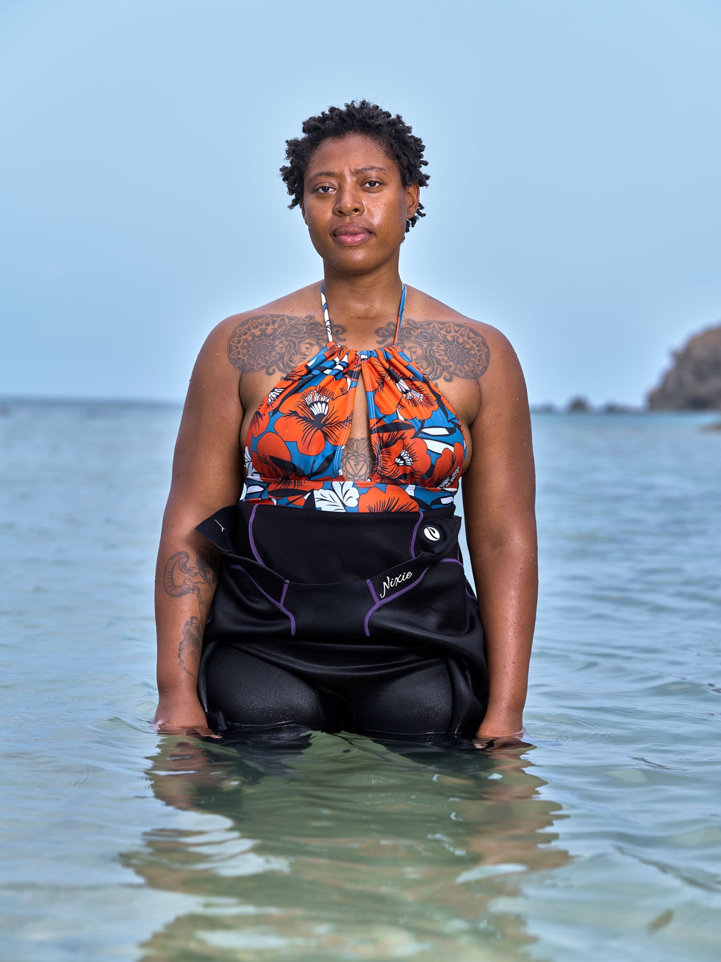 A woman wearing a wet suit and colorful bathing suit top standing in water up to her waist.