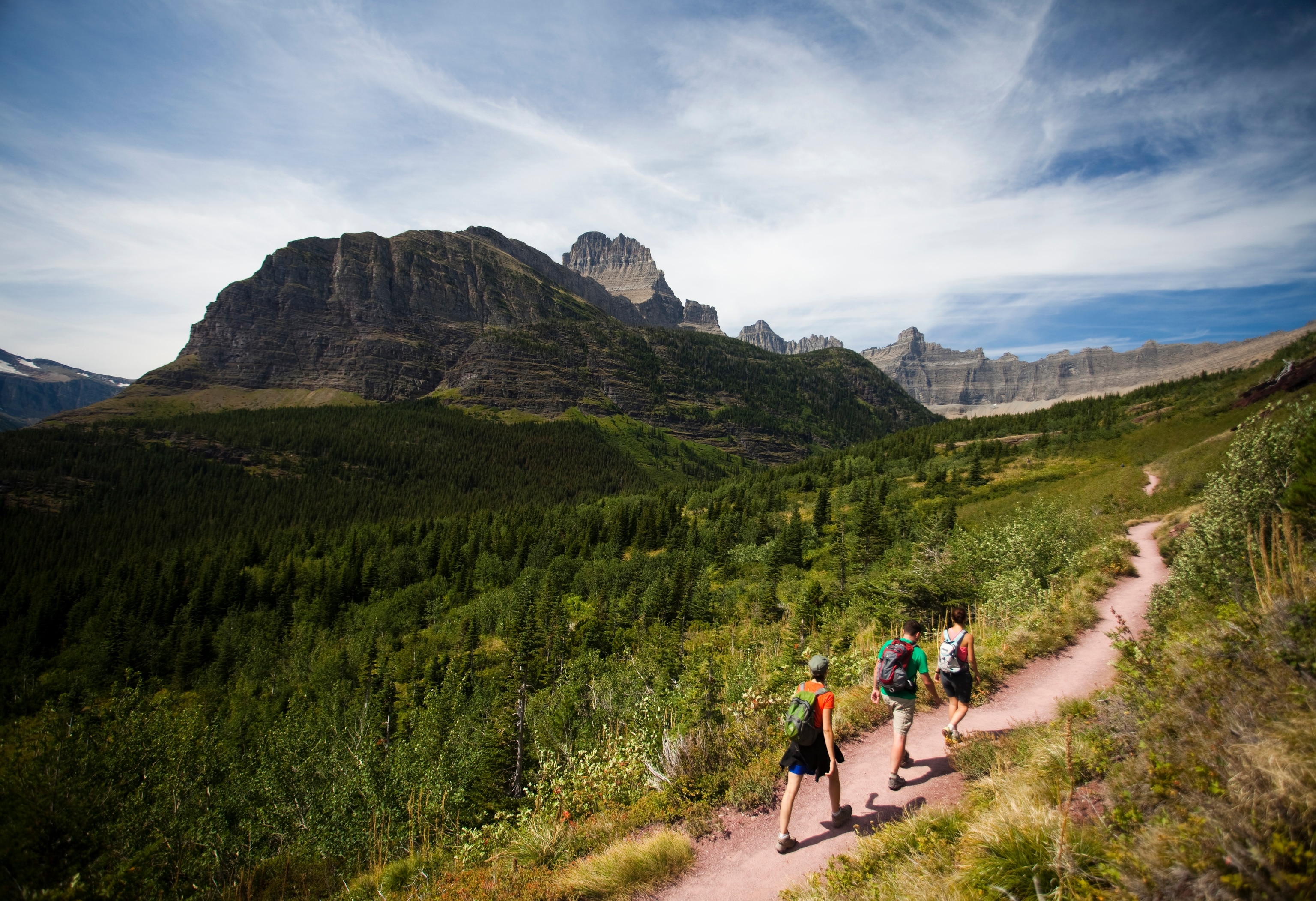 Three hikers walk on a trail with dramatic views of Glacier National Park in the background