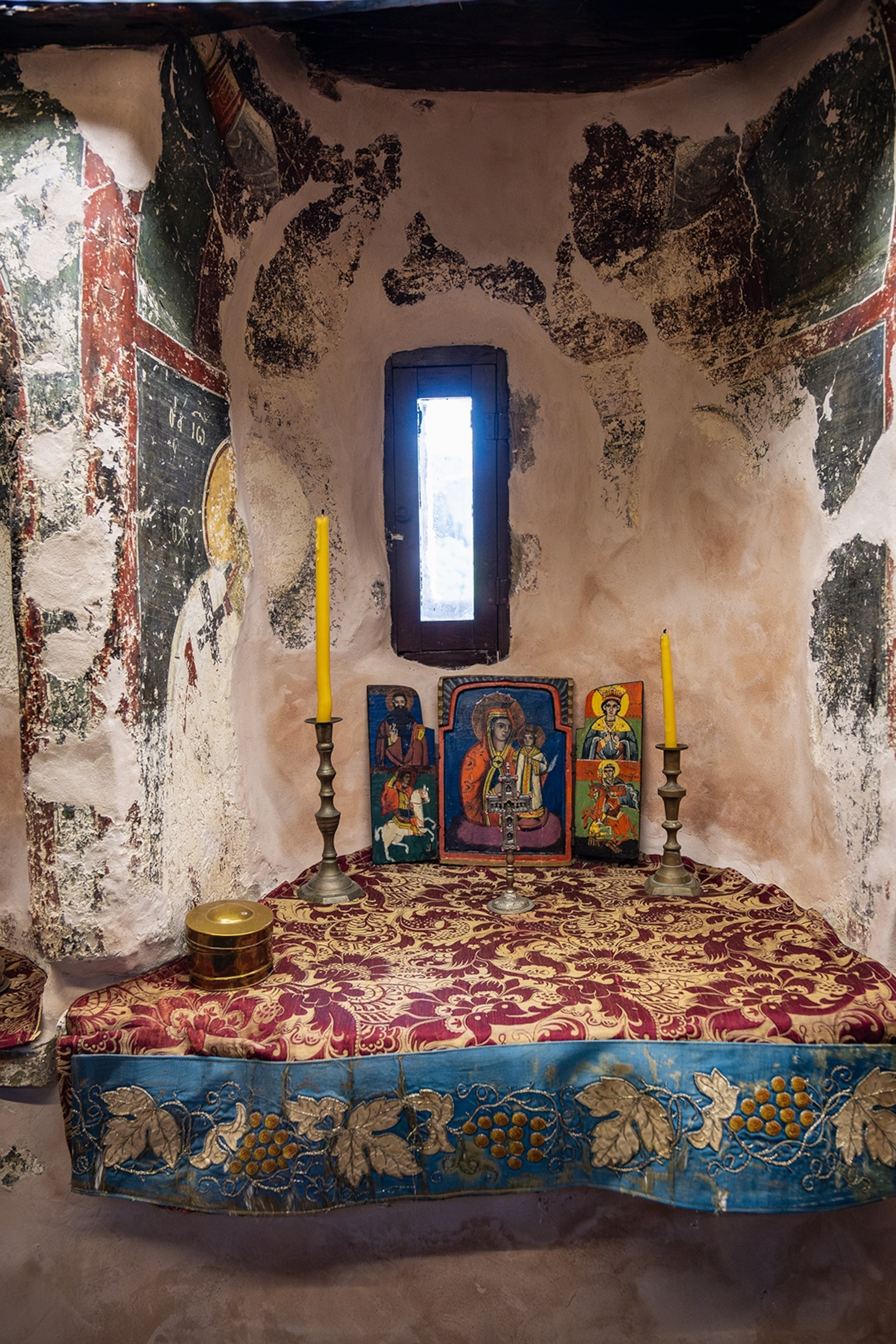 A bed-like altar with a decadent tapestry, tall candles in brass candle holders and religious paintings in a weathered stone room.