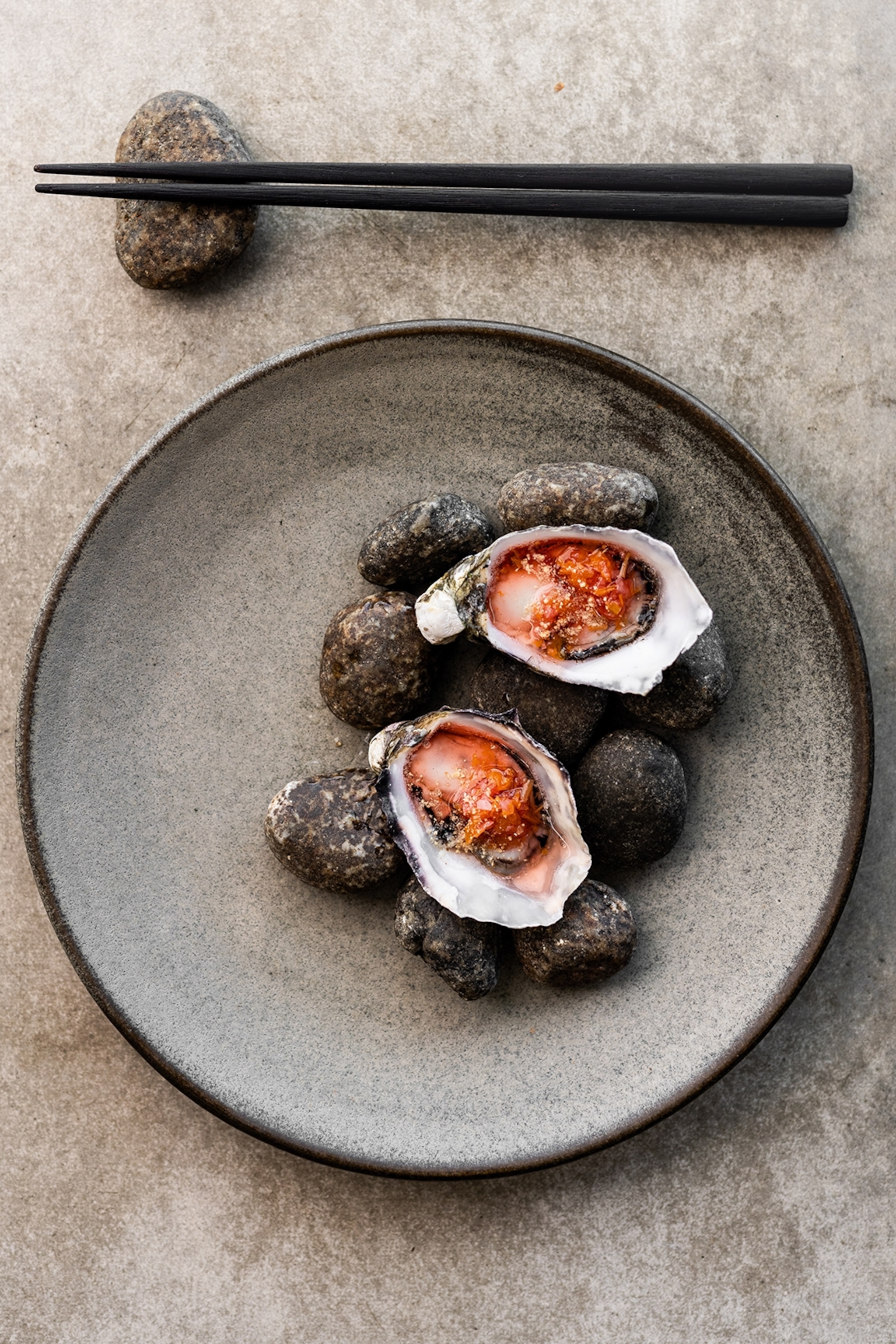 A plate of two oysters on a bed of rocks, with a zingy sauce and a pair of chopsticks on the side.