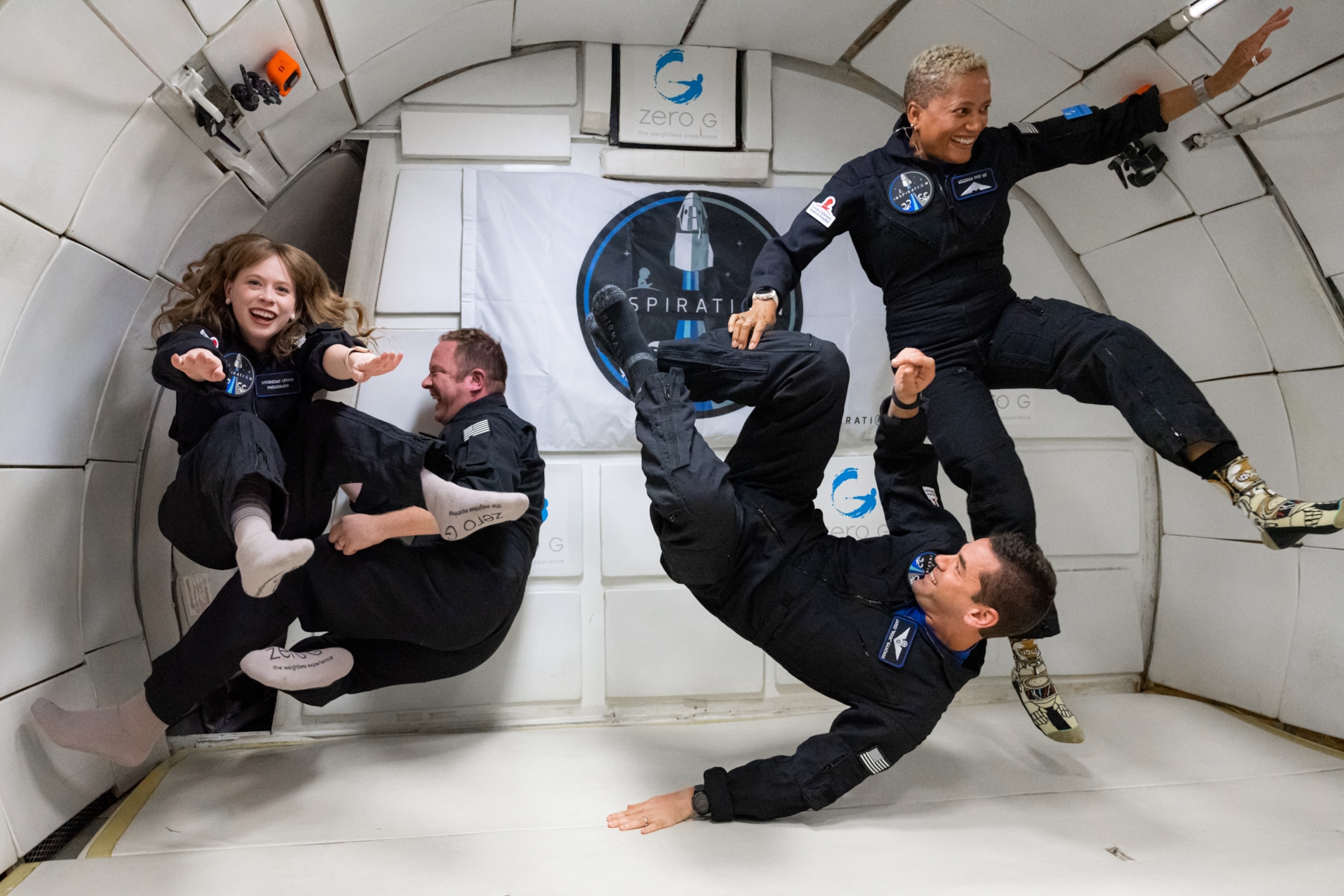 The 4 crew members of Inspiration4, inside the cabin of a Zero-G airplane. The four crew members are in various states of tumbling through the air as they experience weightlessness.