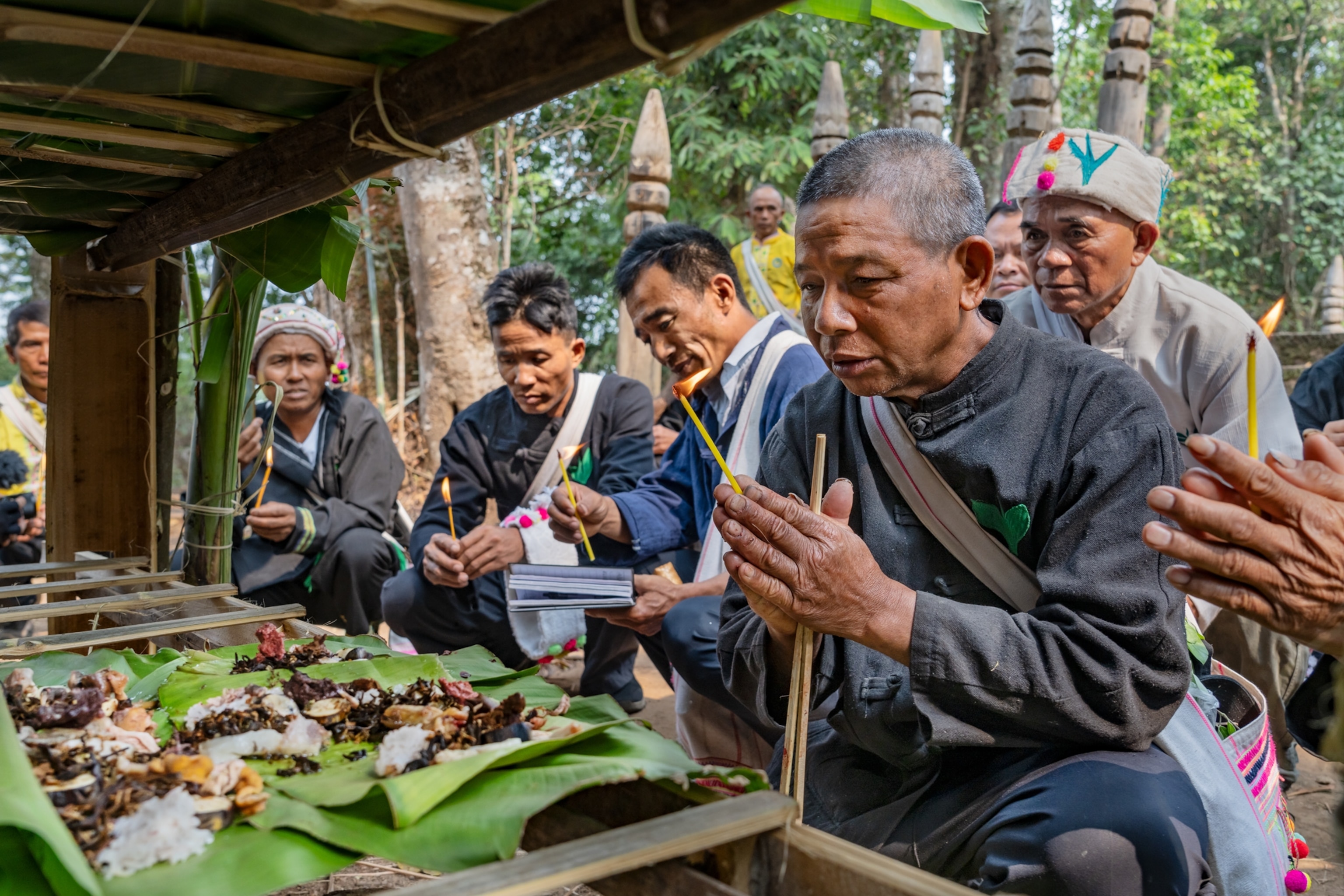 Eight men holding candlelights pray in front of a food offering.