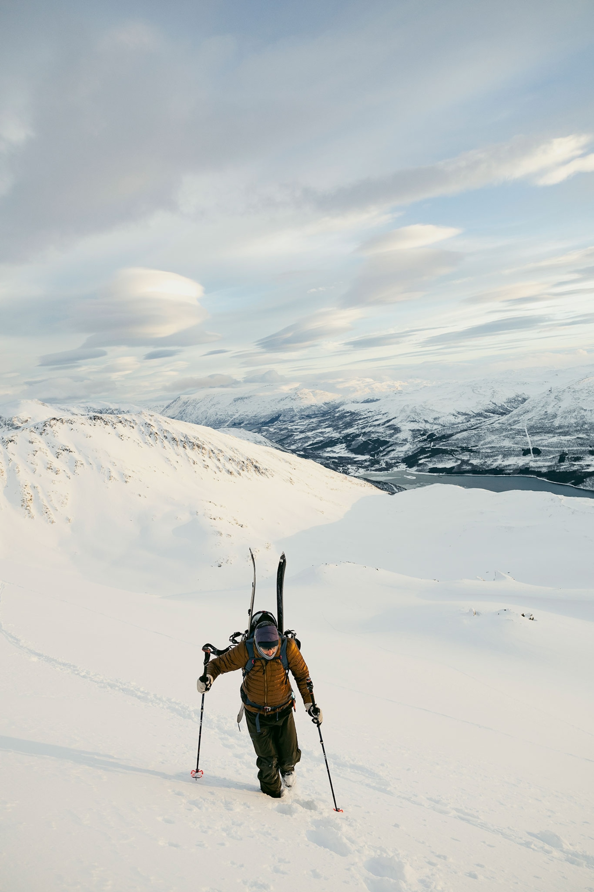 A person skiing up a snow covered mountain. Snow covered mountains and a sky full of clouds fill the frame behind them.