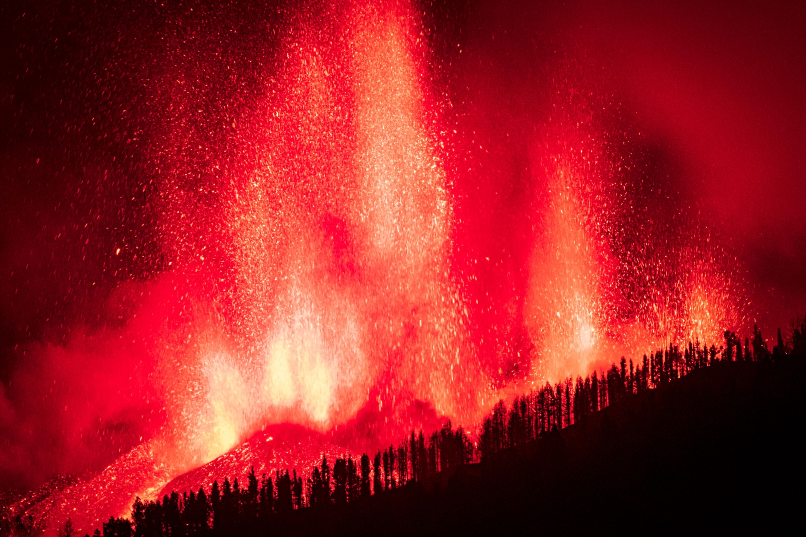 Bright yellow, red, and orange lava shoots up into the dark night sky. The silhouetted trees on the hillside frame the bottom of the image.