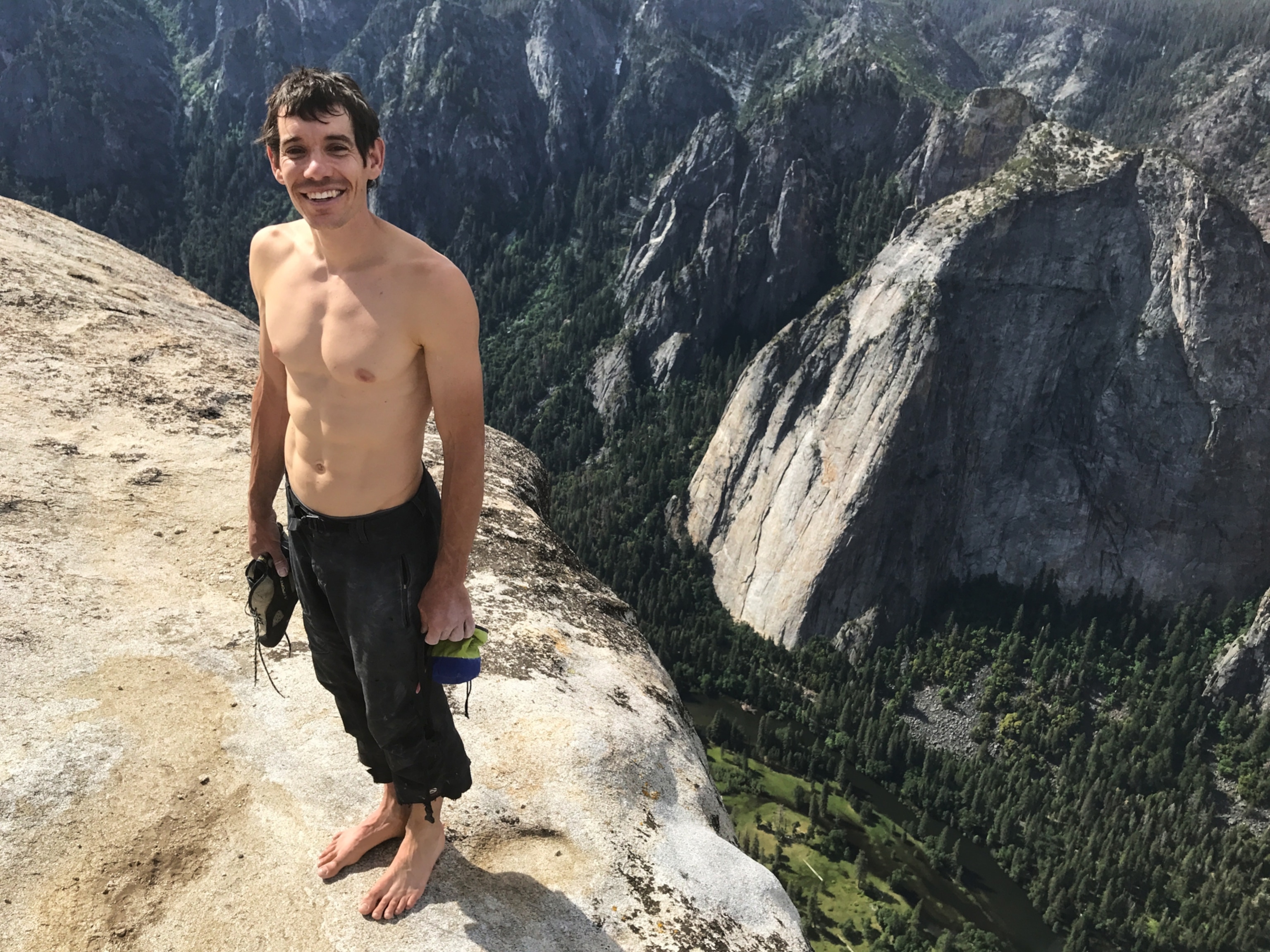 a smiling man at the cliff edge with this shoes in his hands