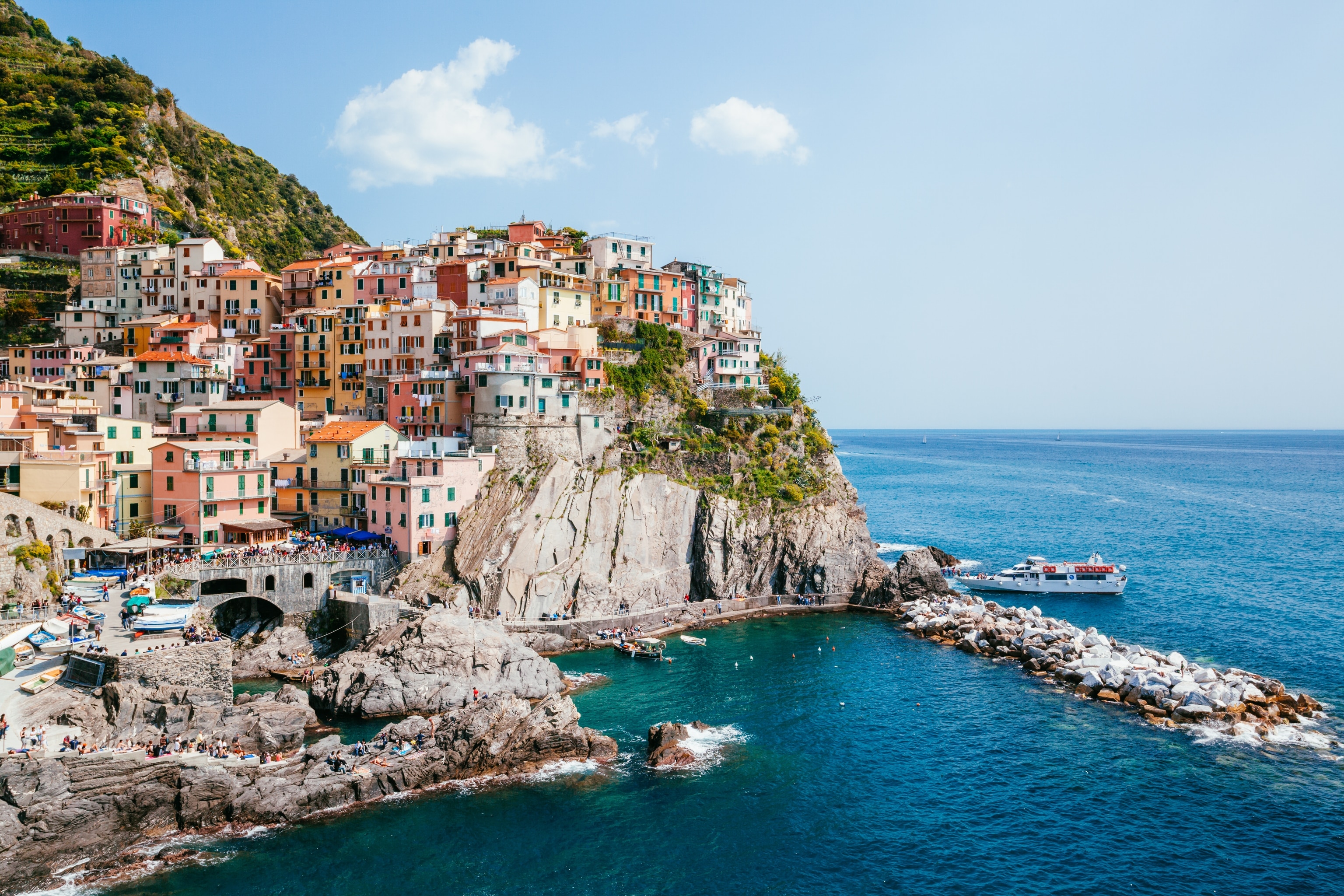 A view of colourful houses atop a cliff in Italy. Below the sea is calm, and a boat is coming in from sea.