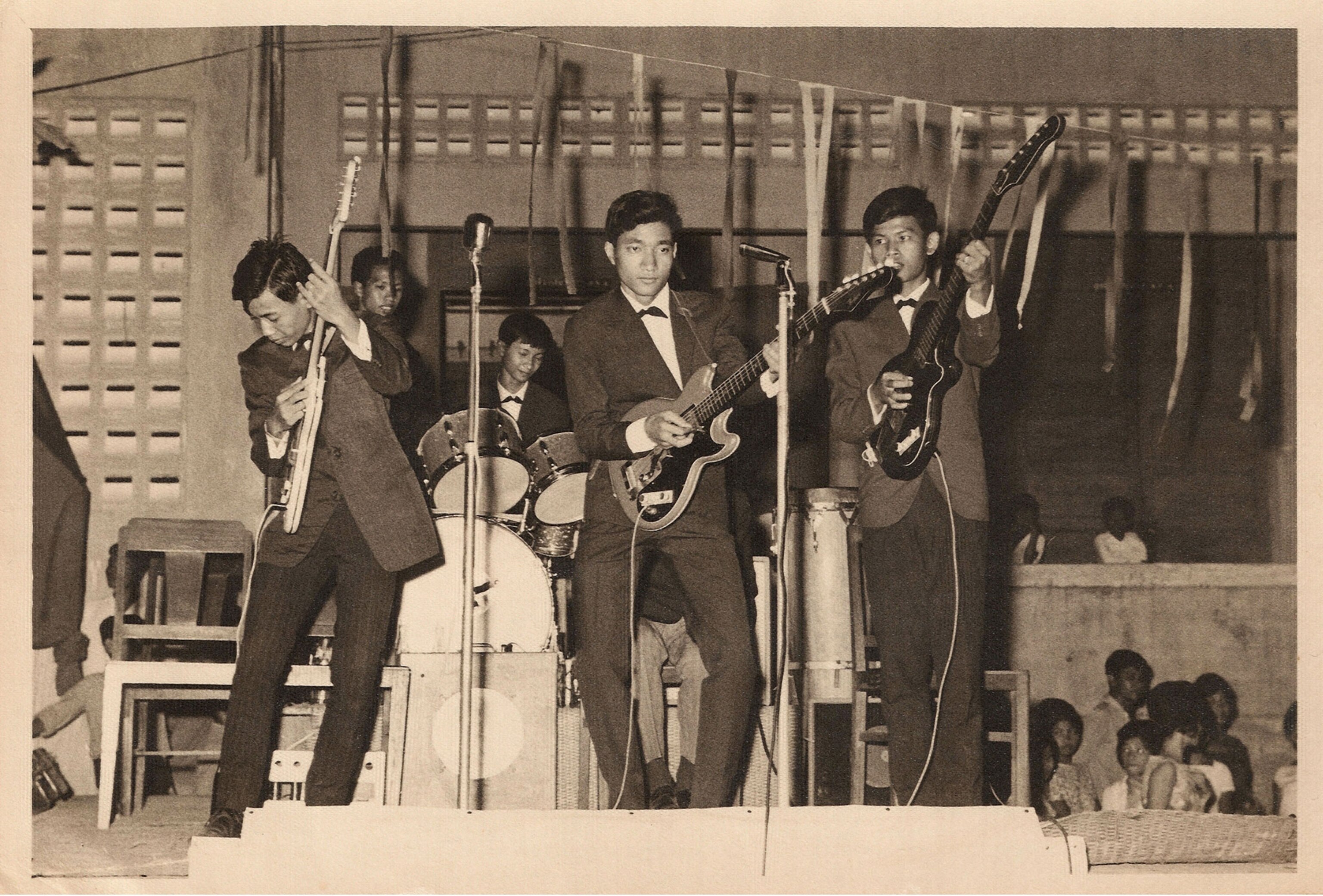 A black and white image of three Cambodian men in a rock band playing guitar on stage.