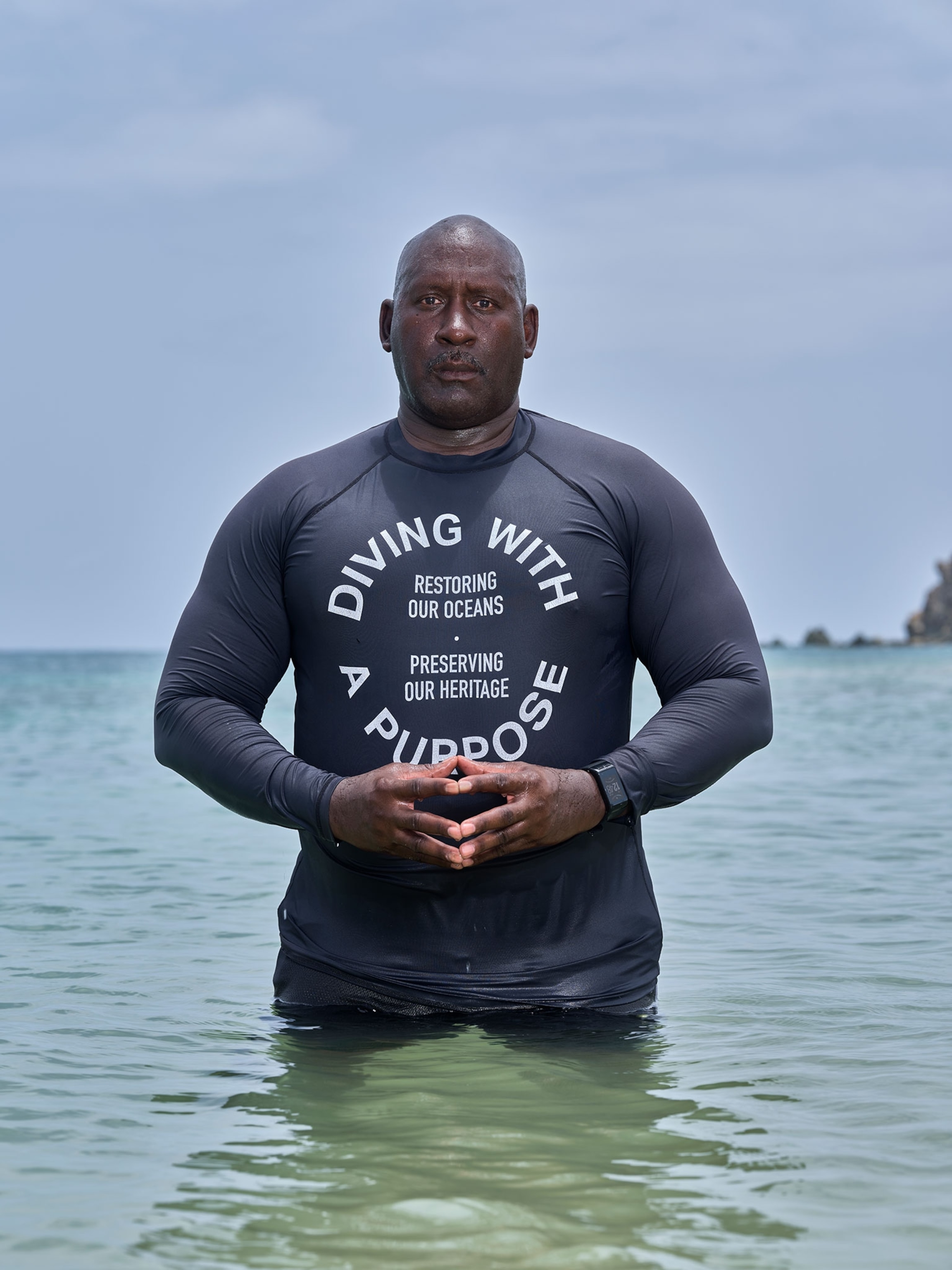 A man in a wet suit that says "Diving with a Purpose" standing in water up to his waist.