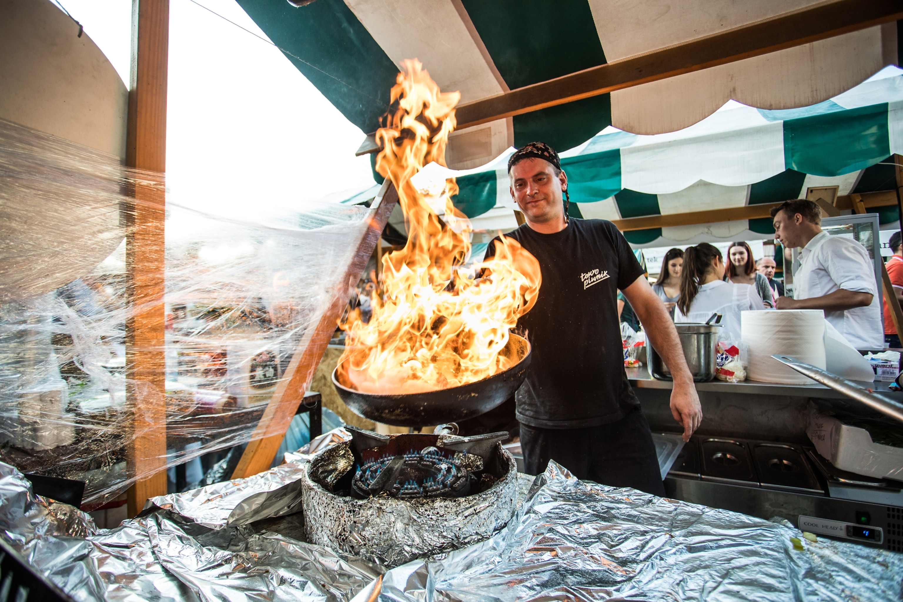 A chef holds a large wok full of flames over a burner in an open-air food market stall, where he cooks for waiting patrons.