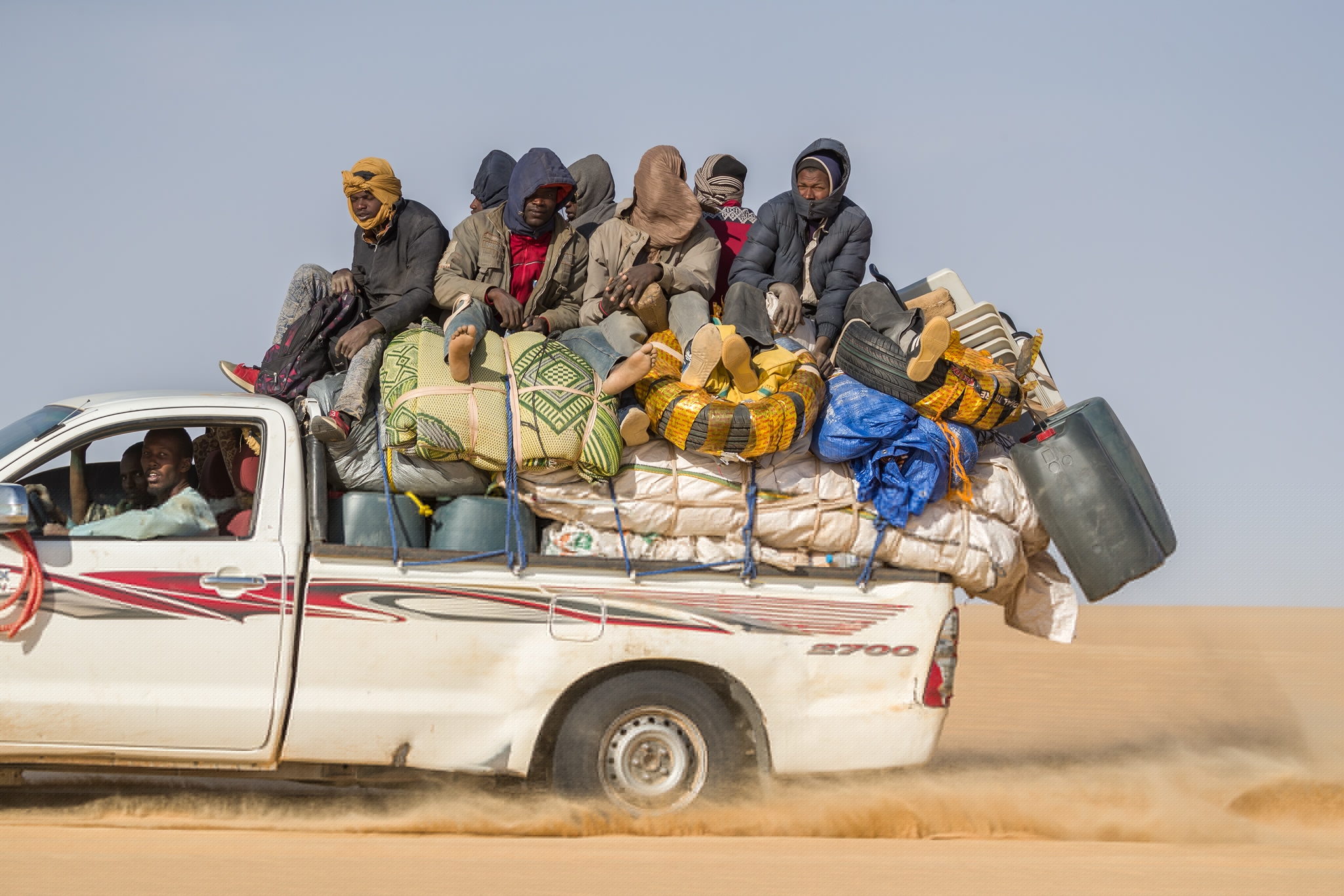 young men sitting on top of goods in a white pick-up