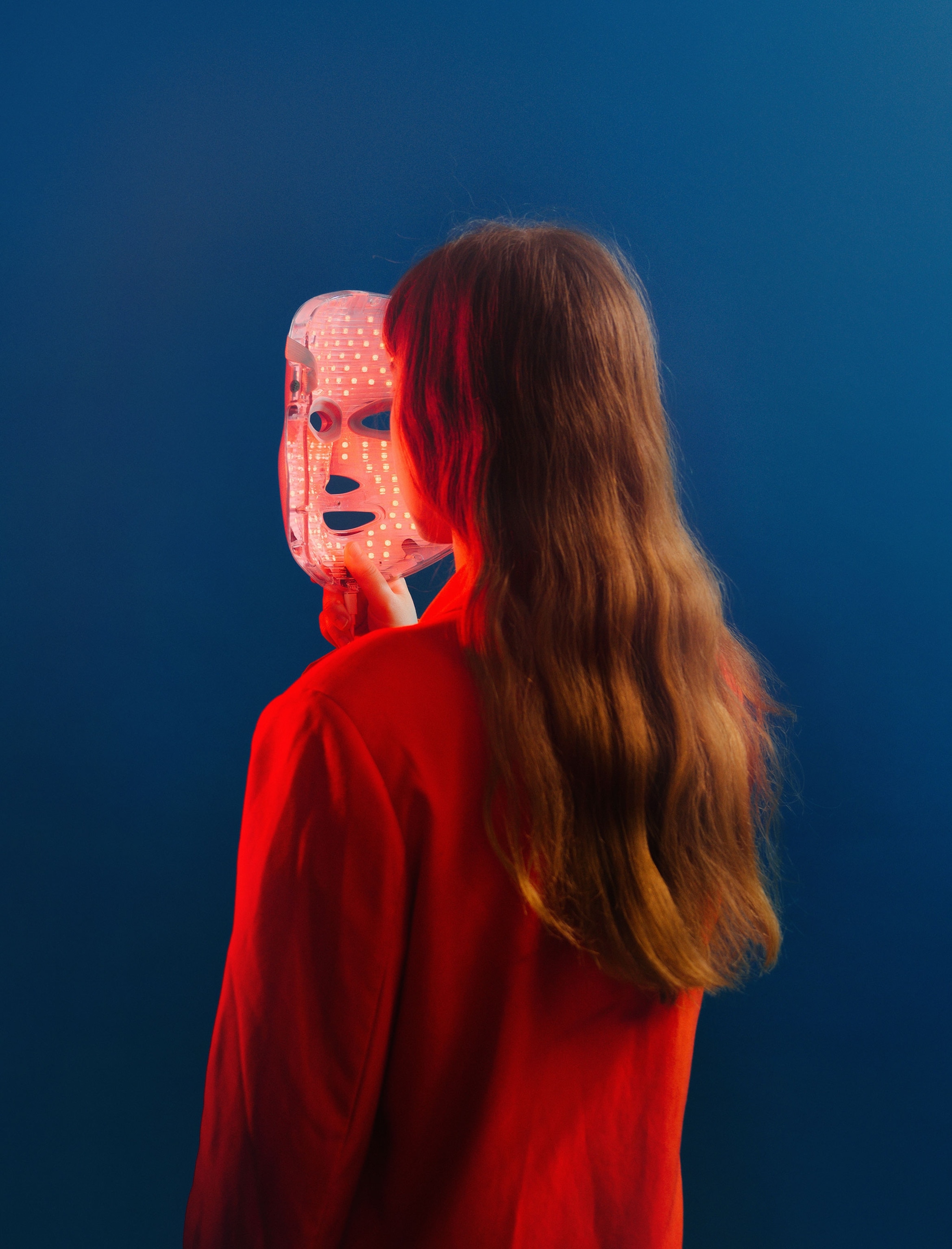 A model poses for a portrait with a red light LED mask designed for consumer use in a studio against a blue background