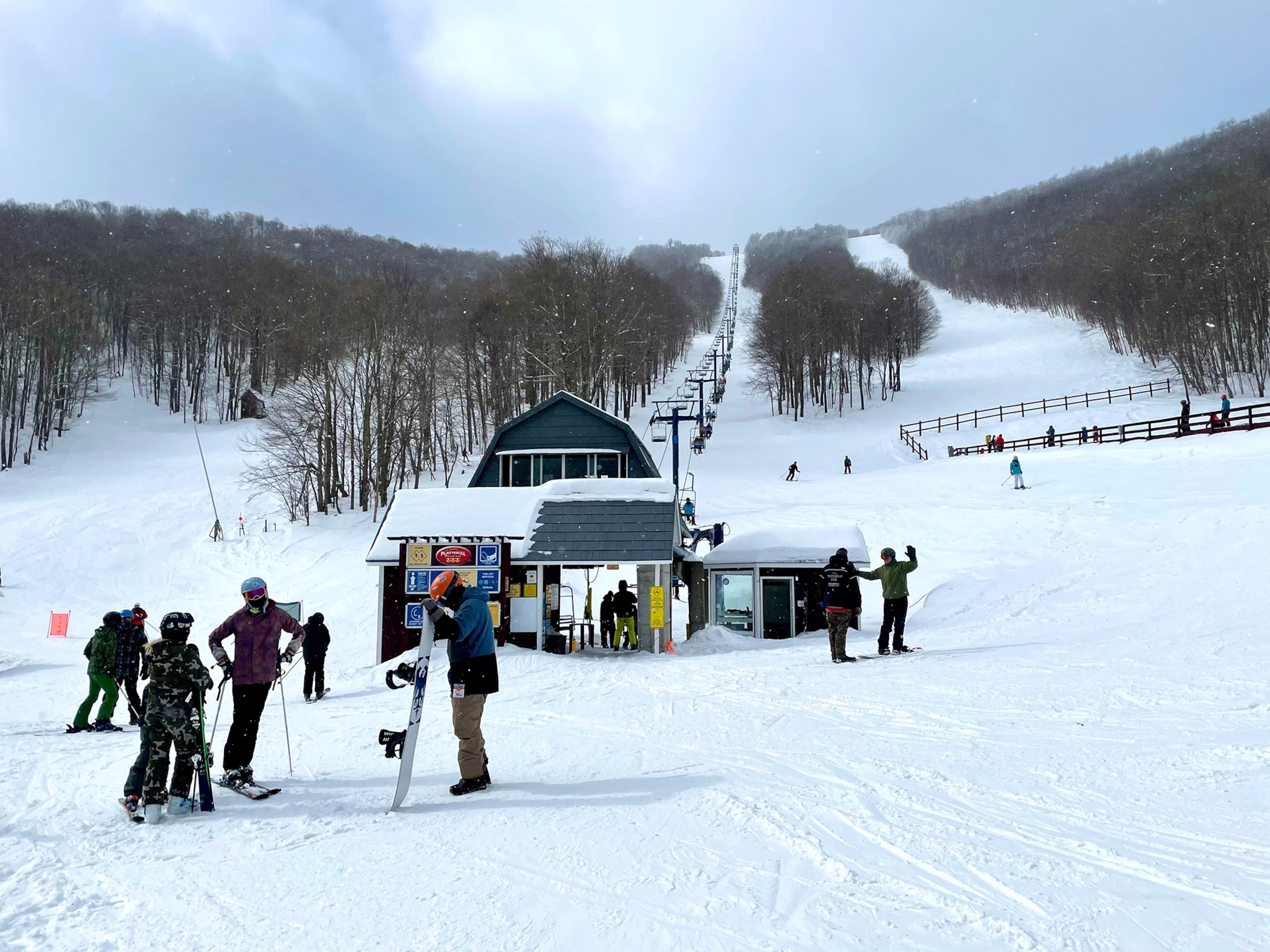 Skiers wait for a chair lift with a building in the background.