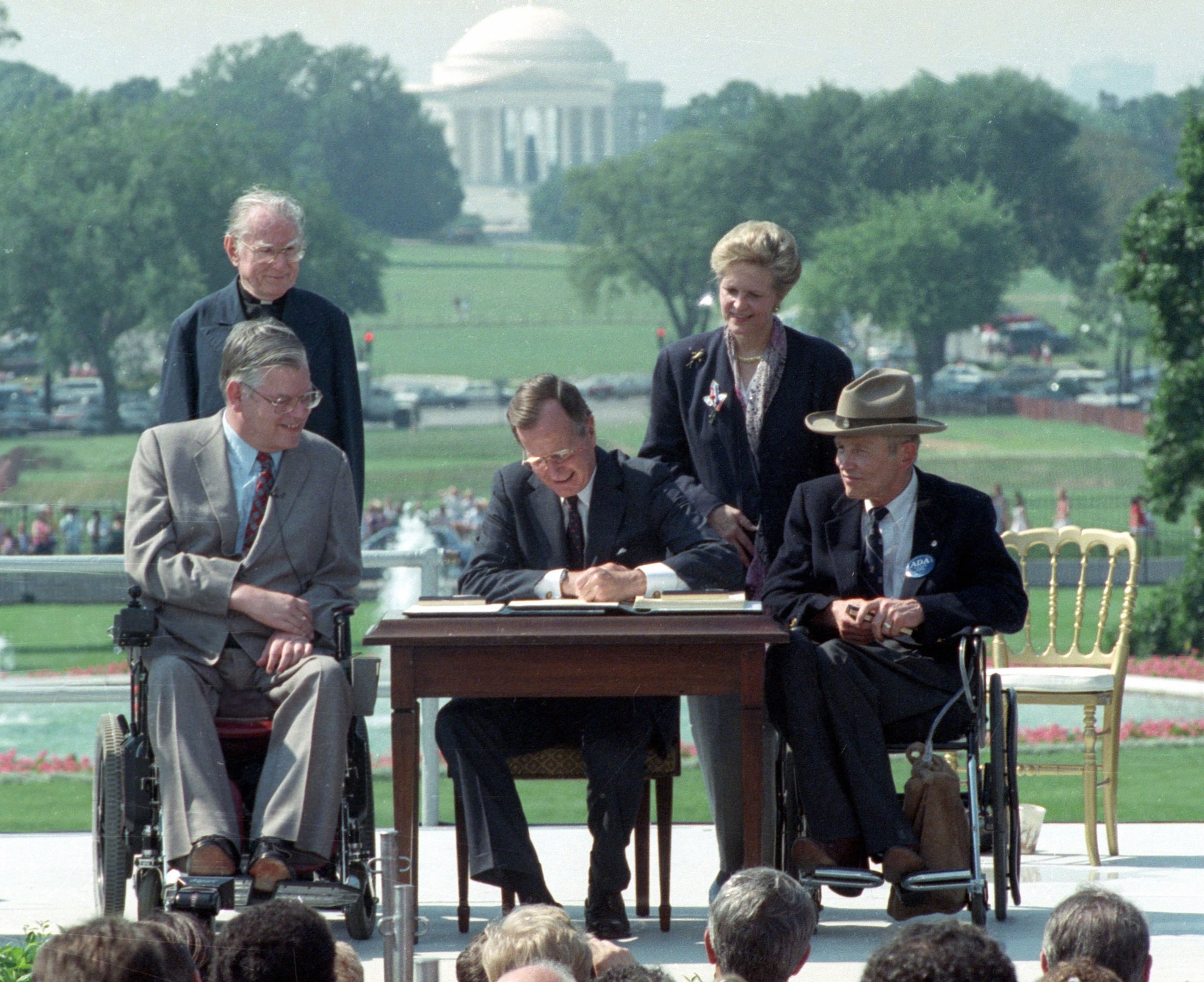 A photograph of a president signing a document outside flanked by two men in wheelchairs