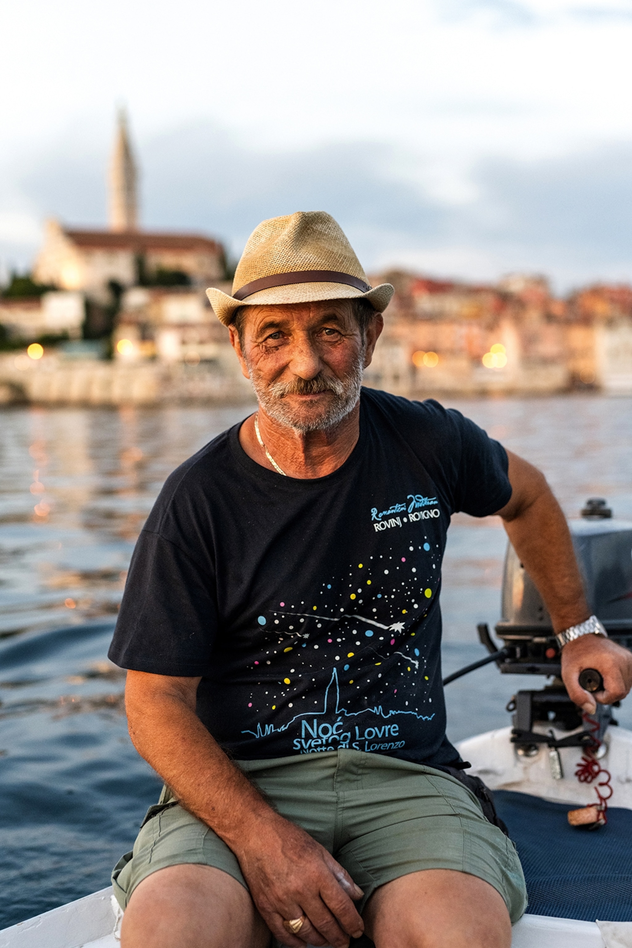 A picture of an elder man wearing a black t-shirt, green shorts and a woven sunhat on a boat with the harbour of Rovinj in the background