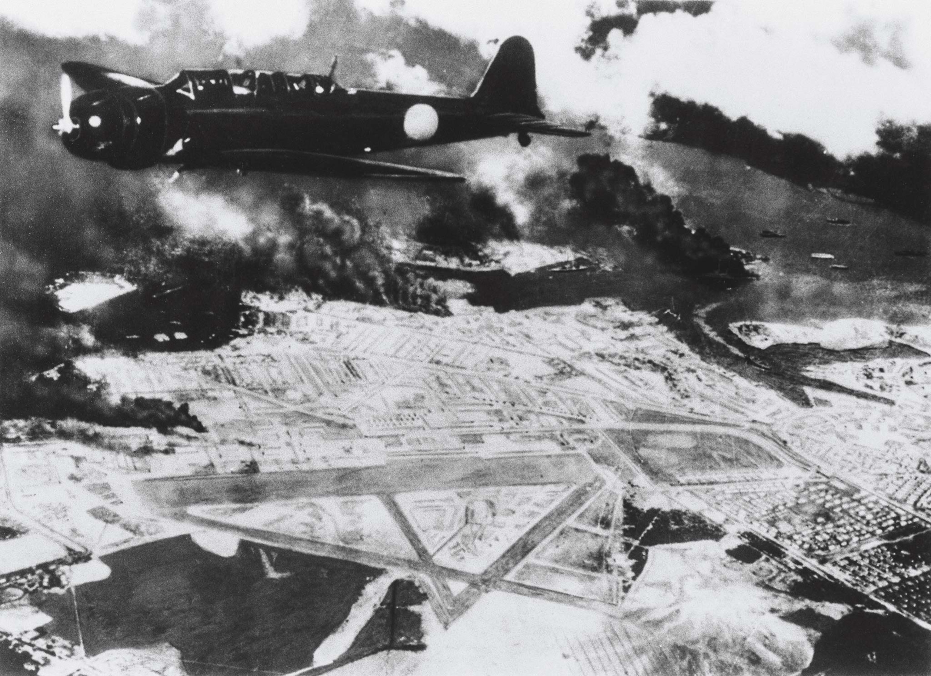 A Japanese bomber flies over an airfield at Pearl Harbor as smoke rises from targeted American warships.