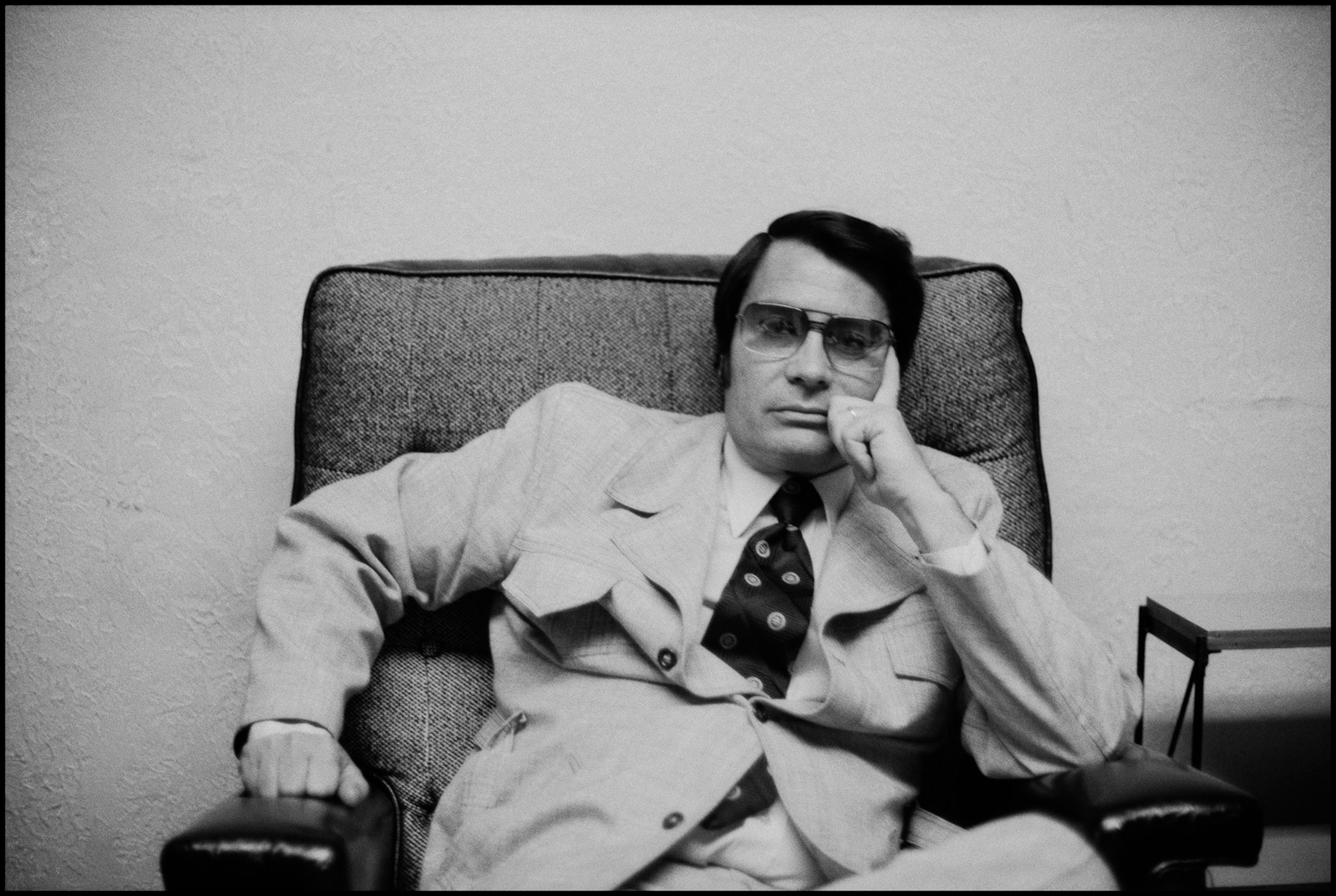 Black and white portrait from the 1970s of man, dressed in a suit, sitting in a chair inside with polarized glasses.