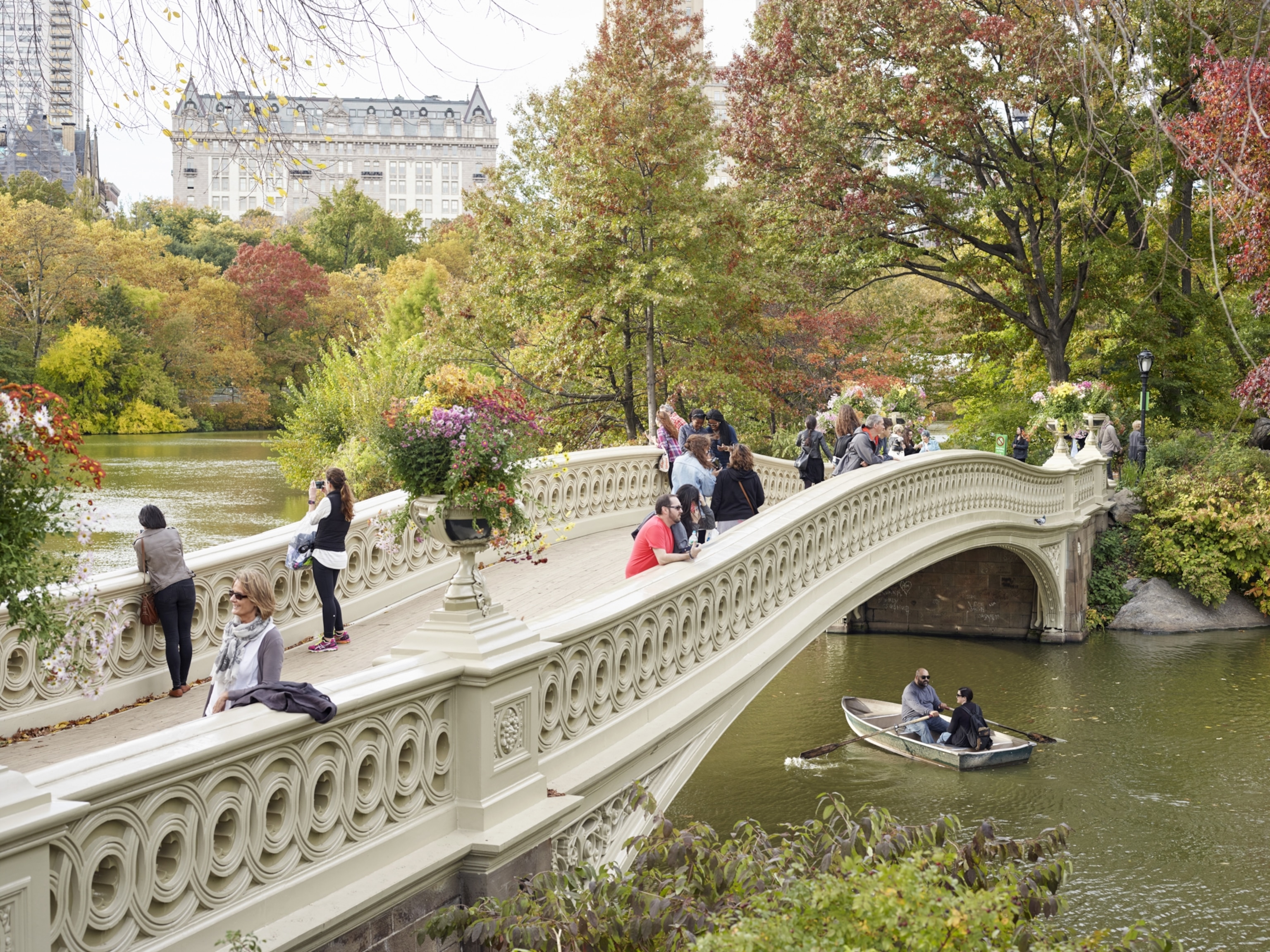 People taking in the sites of Central Park from a white, cast-iron bridge as a couple on a row boat pass beneath.