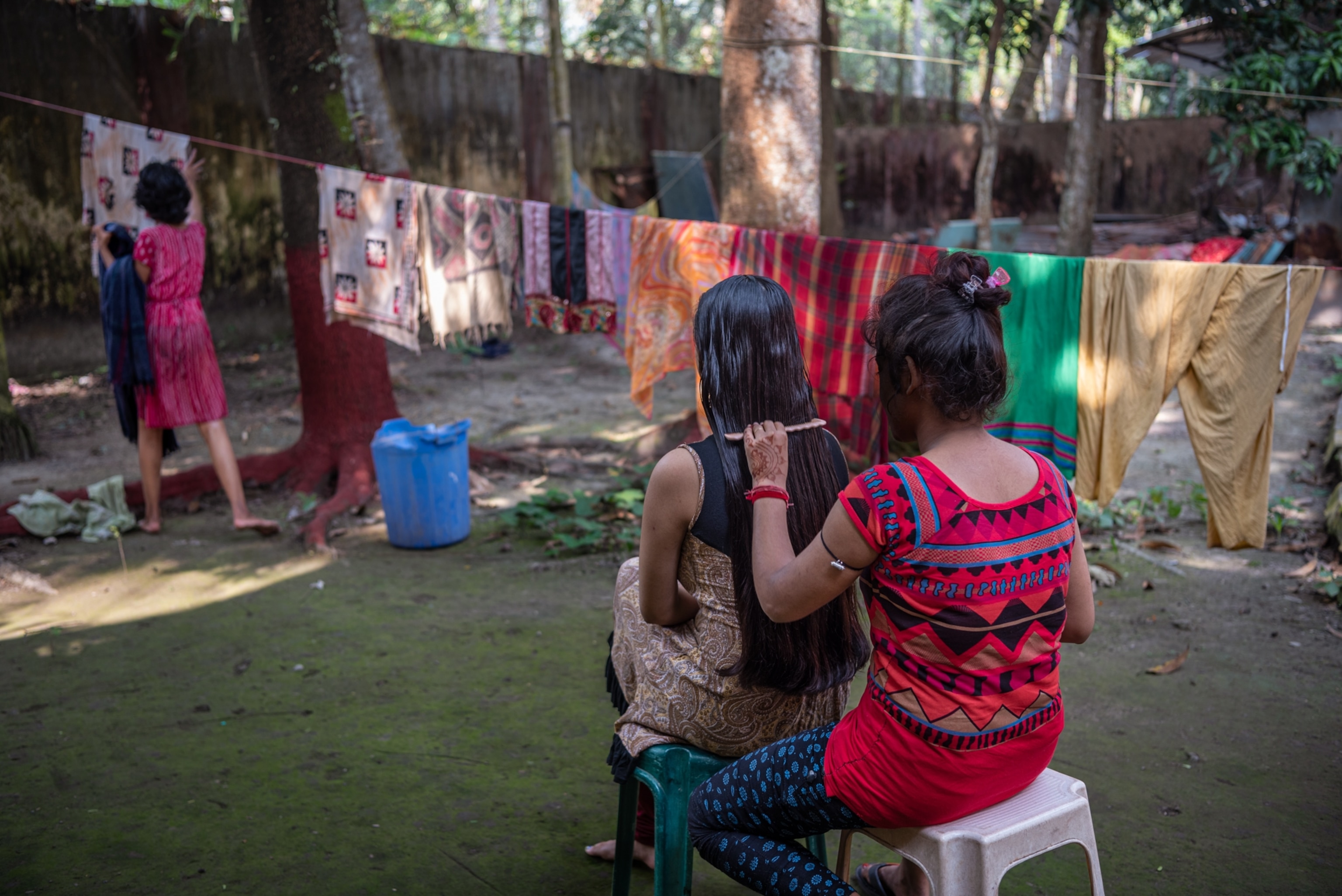 two girls sitting, one brushing the hair of another, while another girl hangs laundry on a line