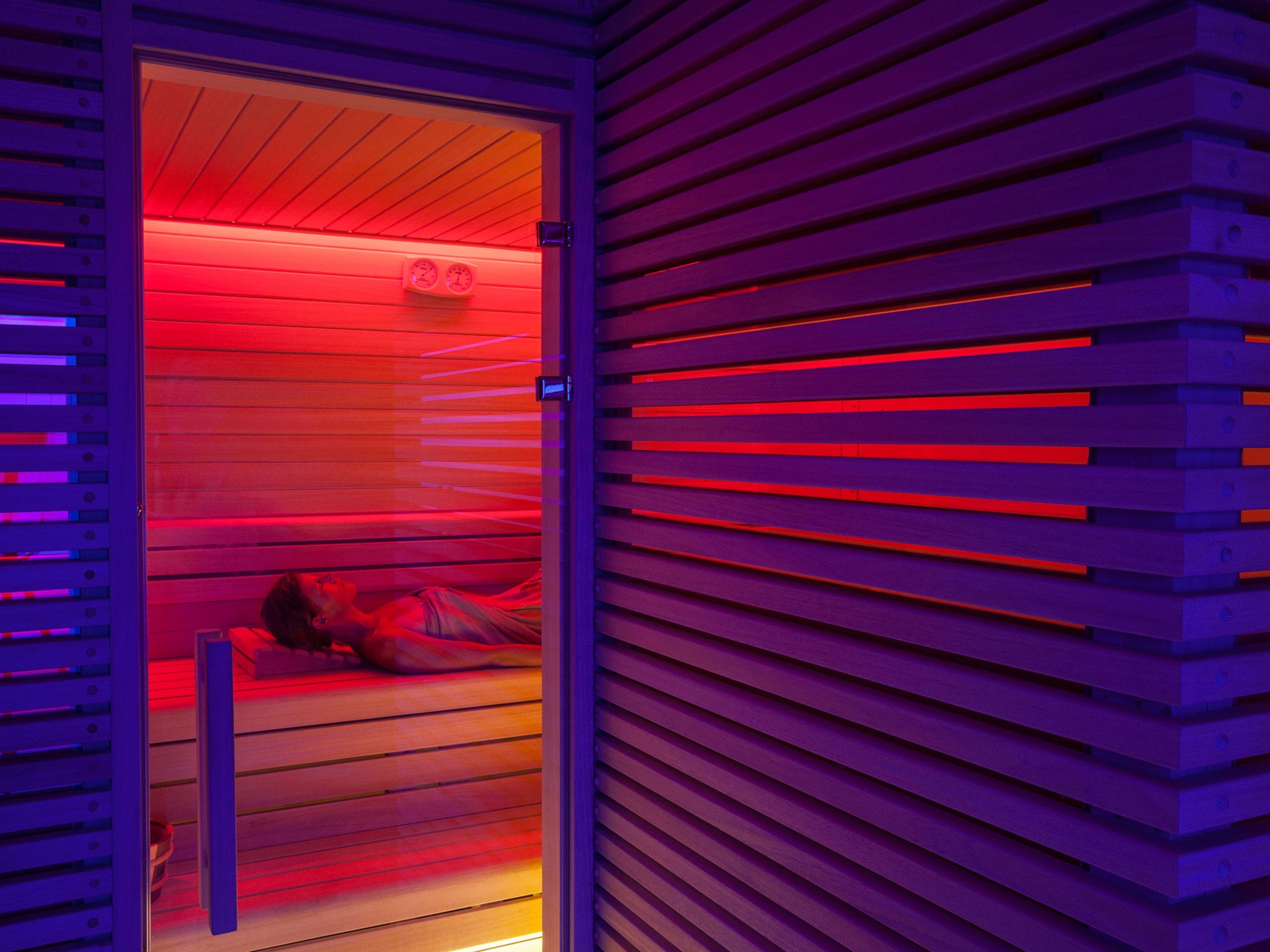 A pink glow is coming from within a sauna where a woman, wrapped in a towel, is lying down on wooden planks. The wood making up the walls outside of the sauna appear purple.