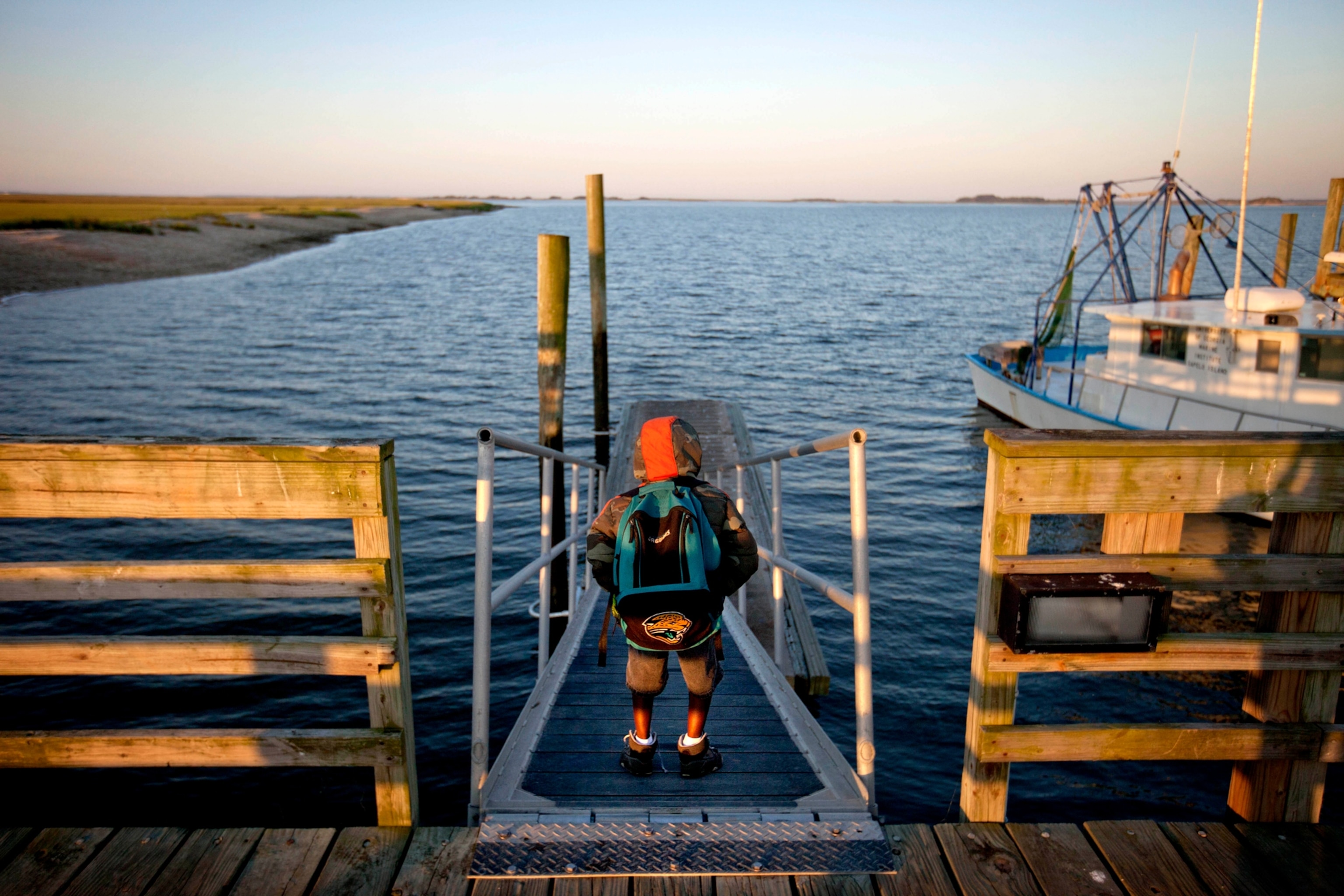 a small child with a rain coat with hood up and large school backpack stands on a dock ramp looking out to empty water