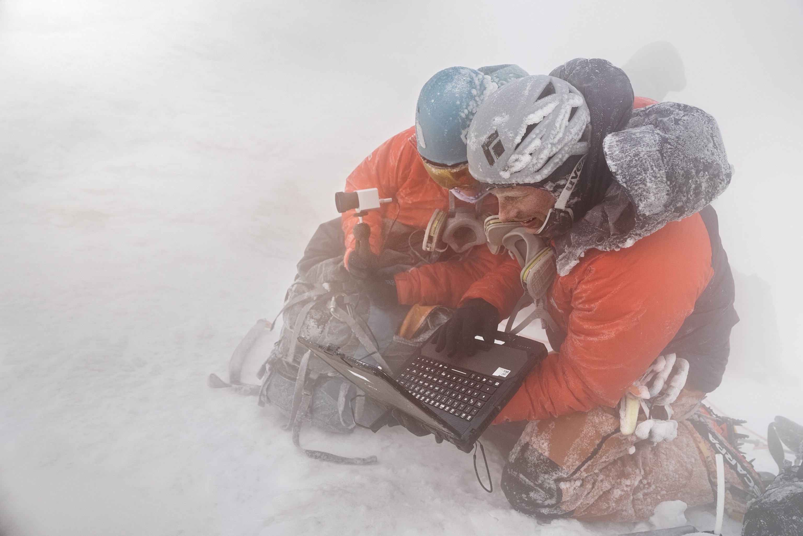 Two people in orange jackets looking into laptop.