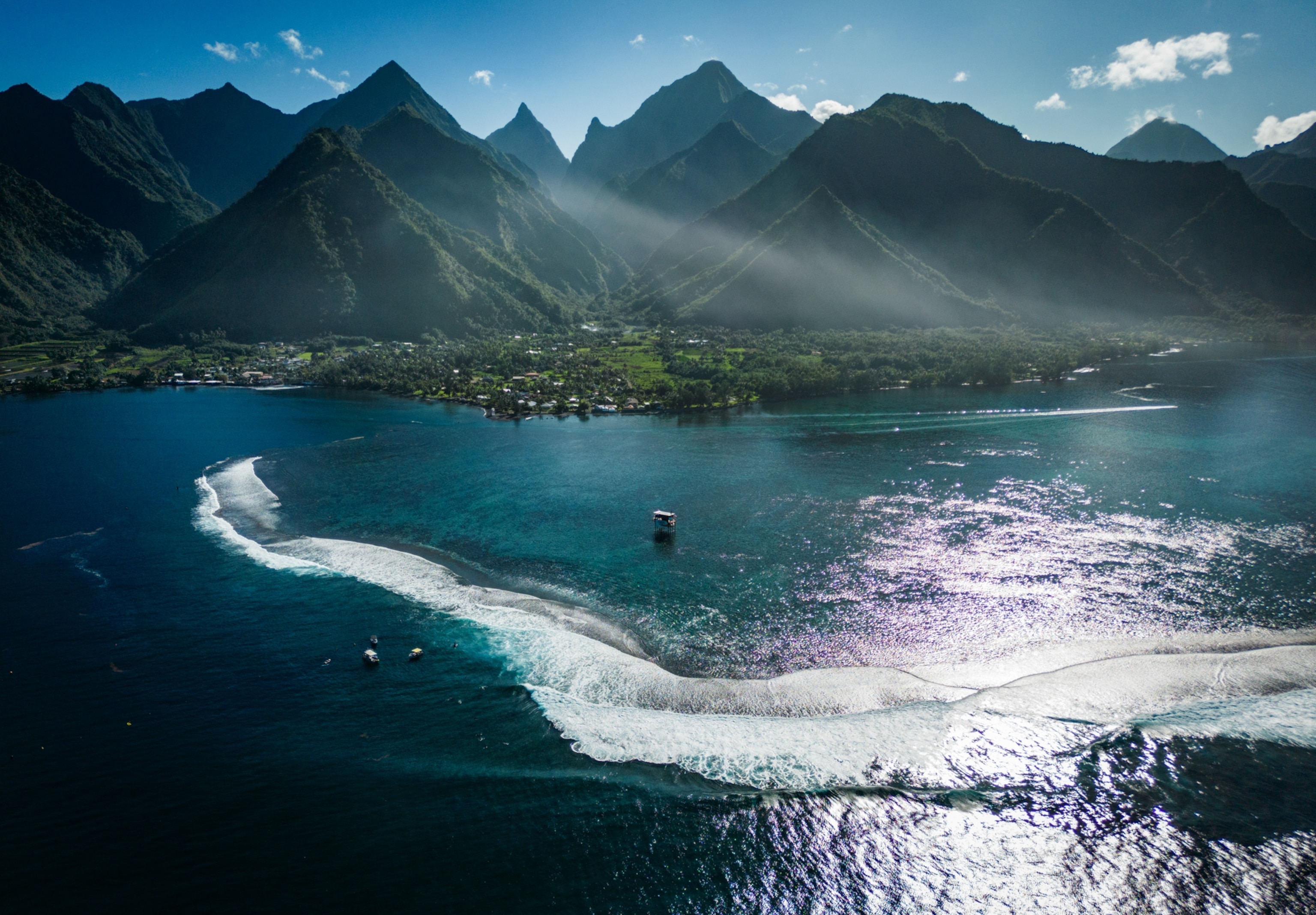 An aerial view of surfers dotting the blue-green waters off the coast of Teahupo'o as a white ribbon of wave rolls towards the coast. The coastline appears lush and green before giving way to dozens of green, pointed mountain peaks.