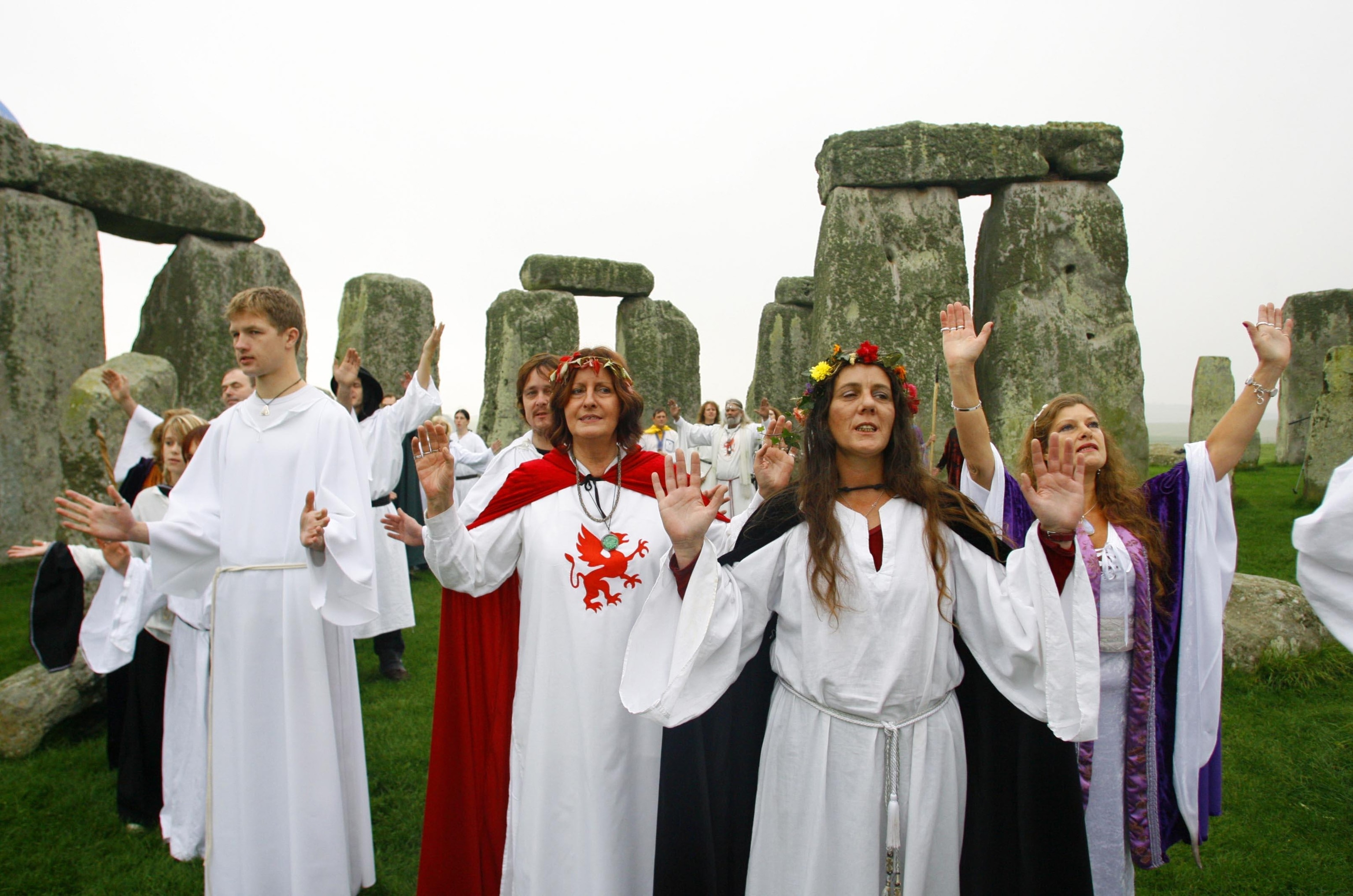 Several white men and women wearing white robes emblazoned with Celtic symbols stand with their hands up, performing a ritual in front of stone megaliths in a grassy field.