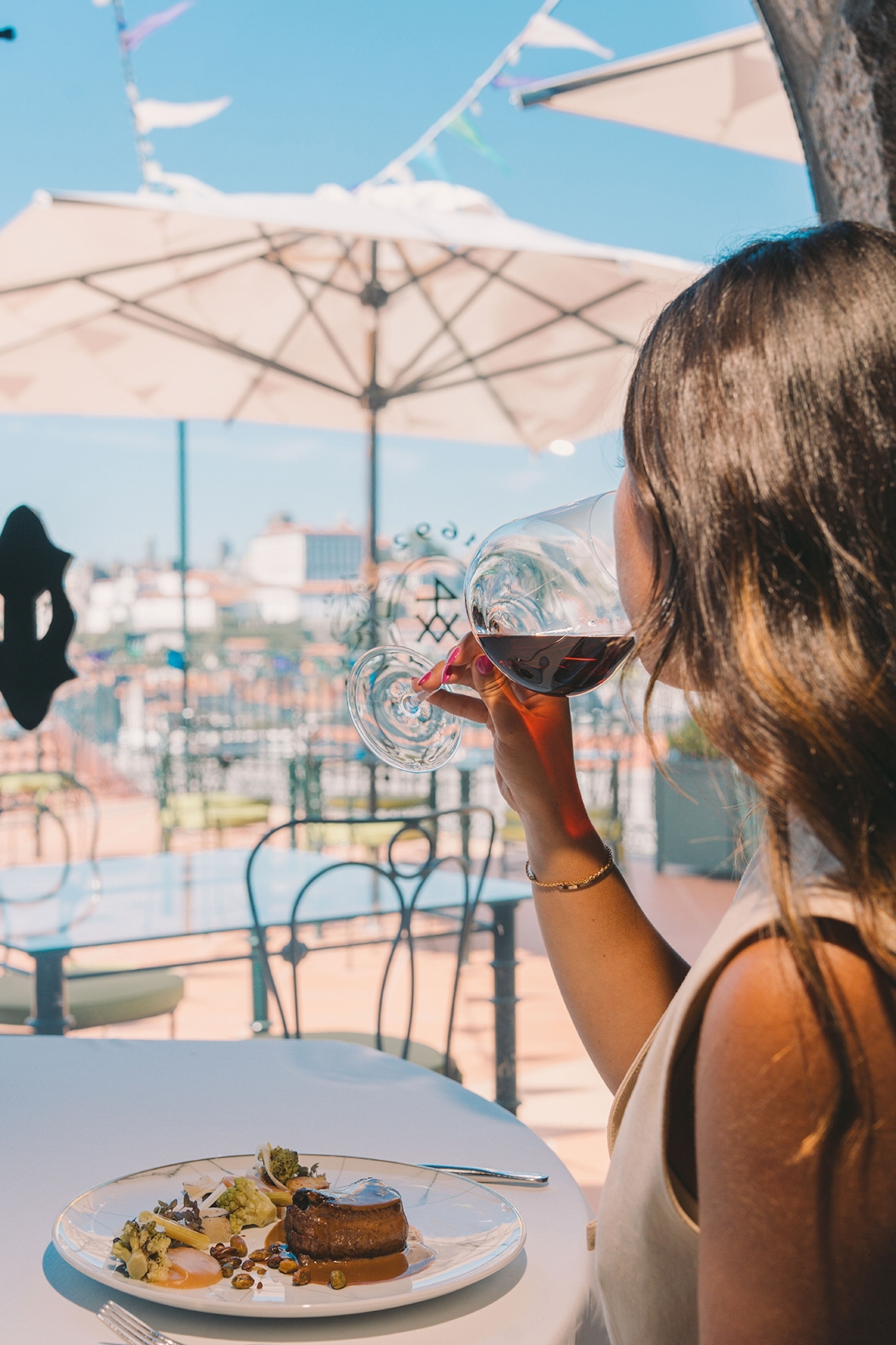 A woman sipping red wine and sat a table with a plate of steak, looking out at the cityscape, shot from behind
