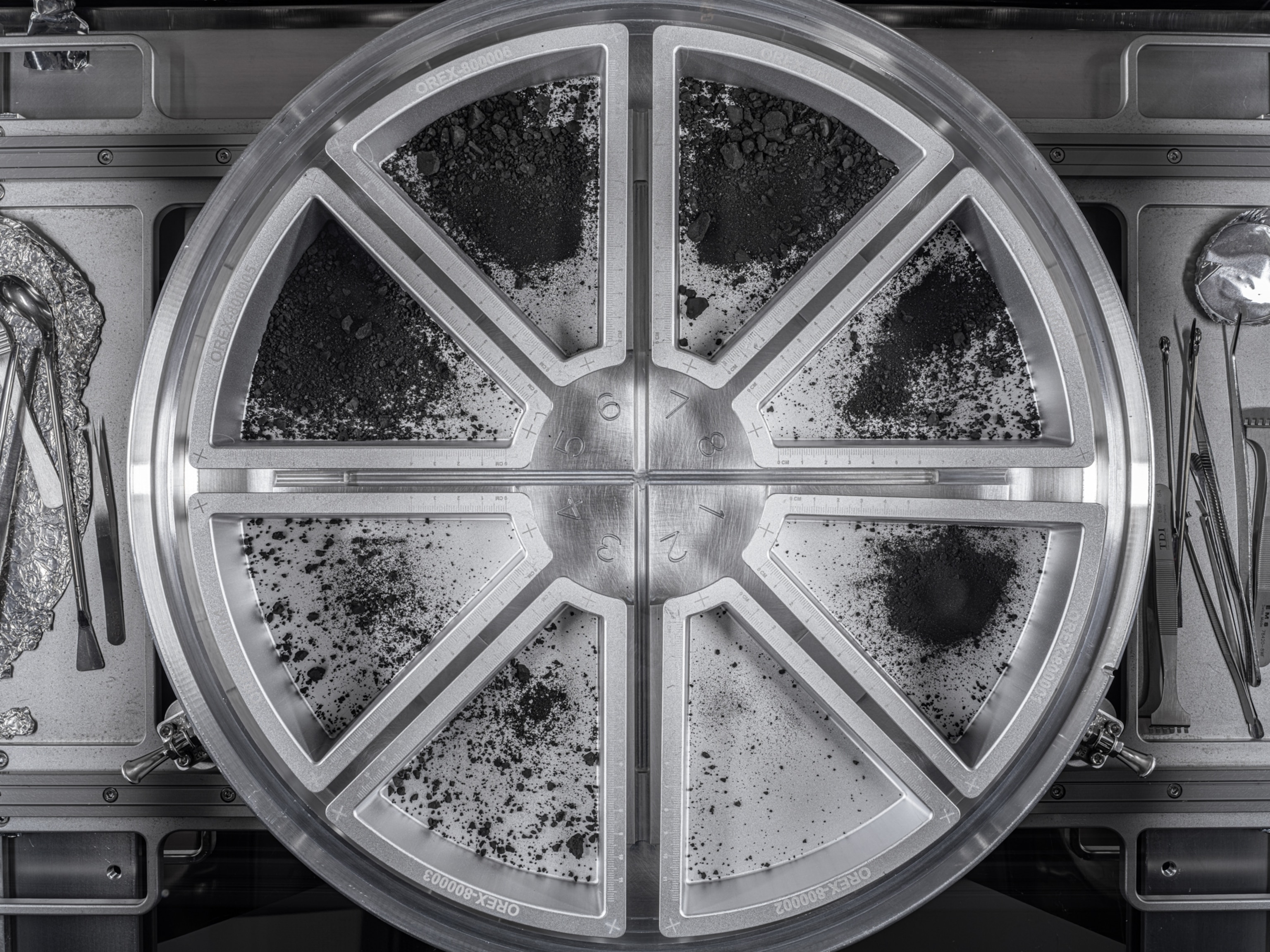 A top down view of a round metal container divided into 8 sections, each of which contains some black sediment and rocks from asteroid Bennu.