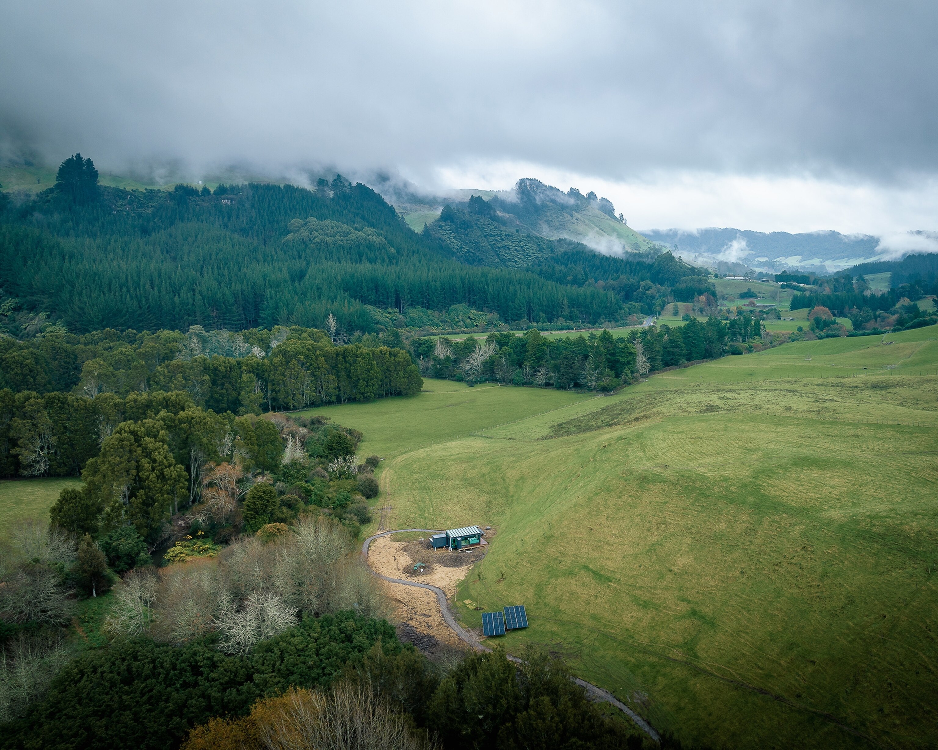 An aerial view of the Pāmu PurePod, a modernist glass and wood cabin surrounded by a forest (left) and open field (right) in Rotorua, New Zealand.