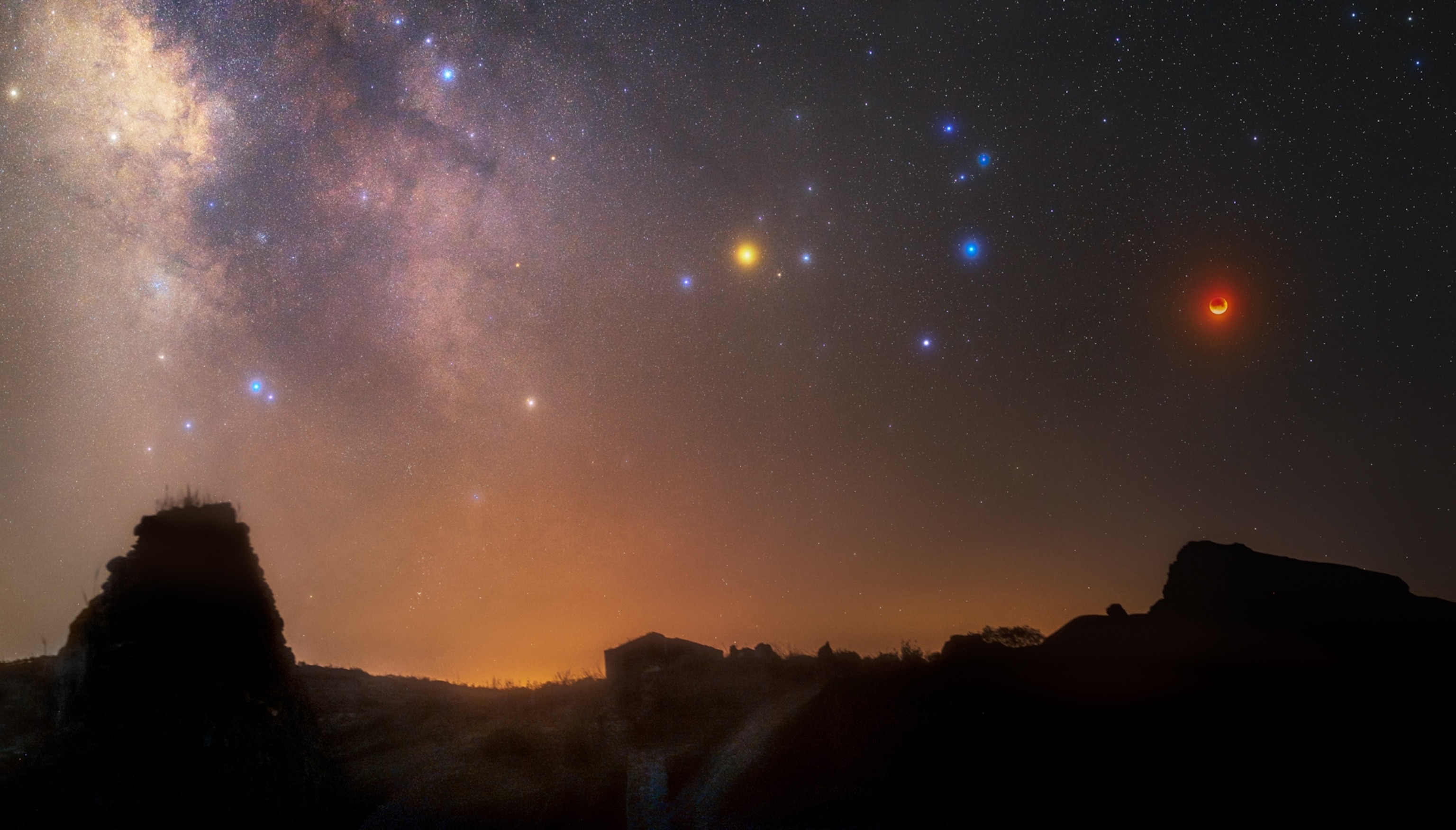 A wide scene of a mountain range beneath the milk way. The moon is small, but visibly red. Another yellow-orange bright source of light is seen in the middle of the starry night sky.