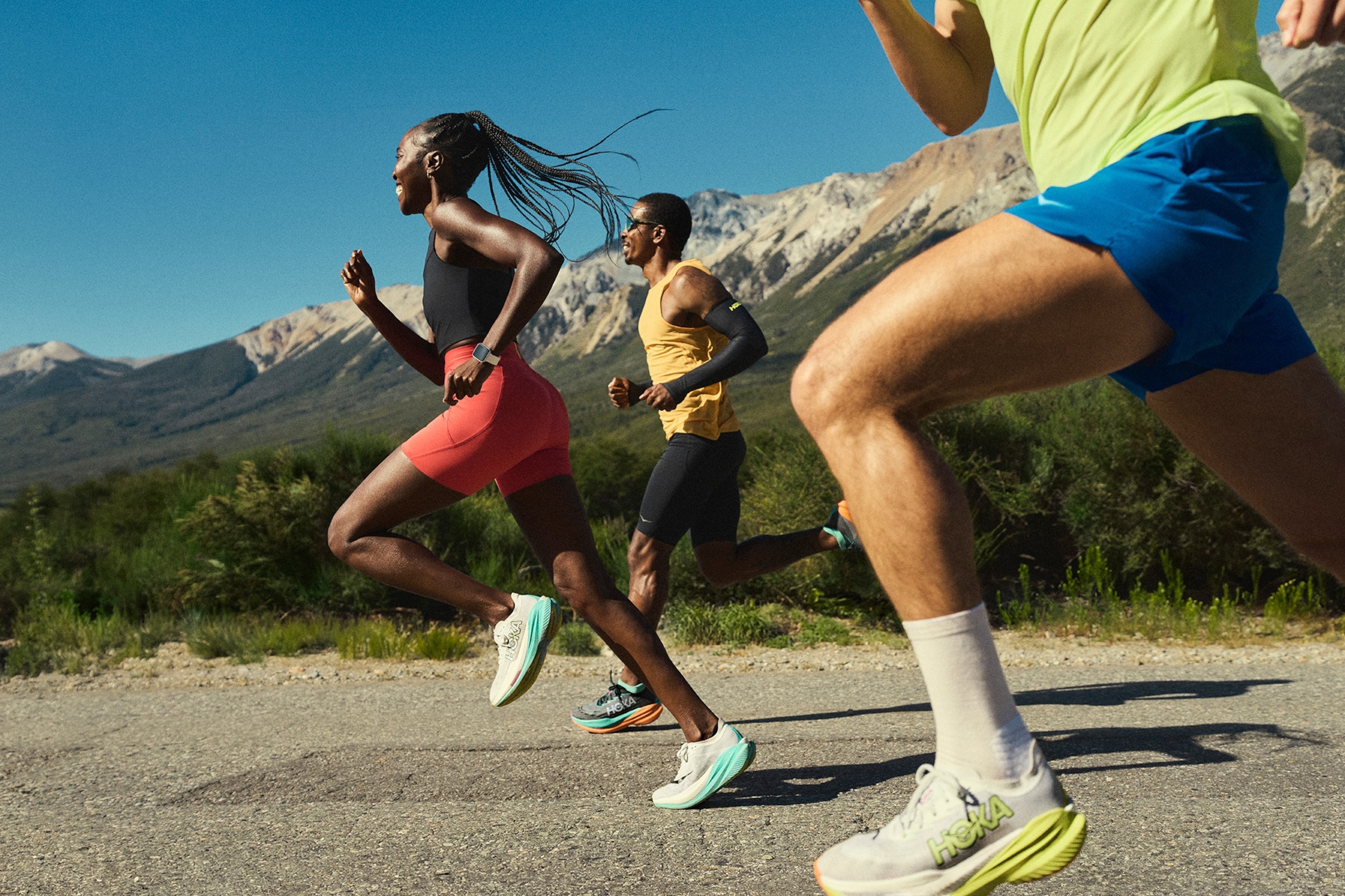 Three runners wearing Hoka shoes with mountains in the background.