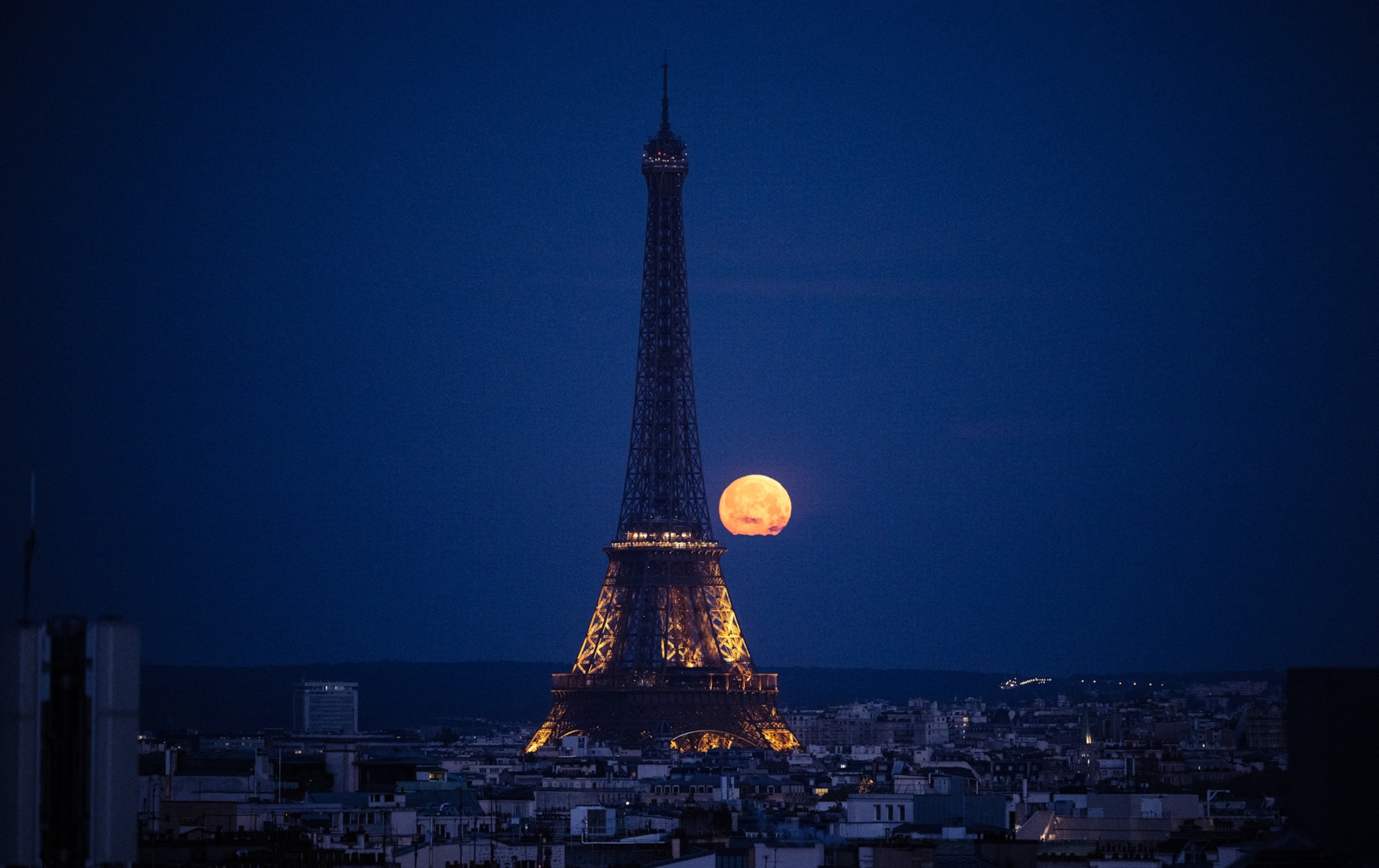 A full moon glows orange against a deep blue evening sky, as the moon rises behind the Eiffel Tower.