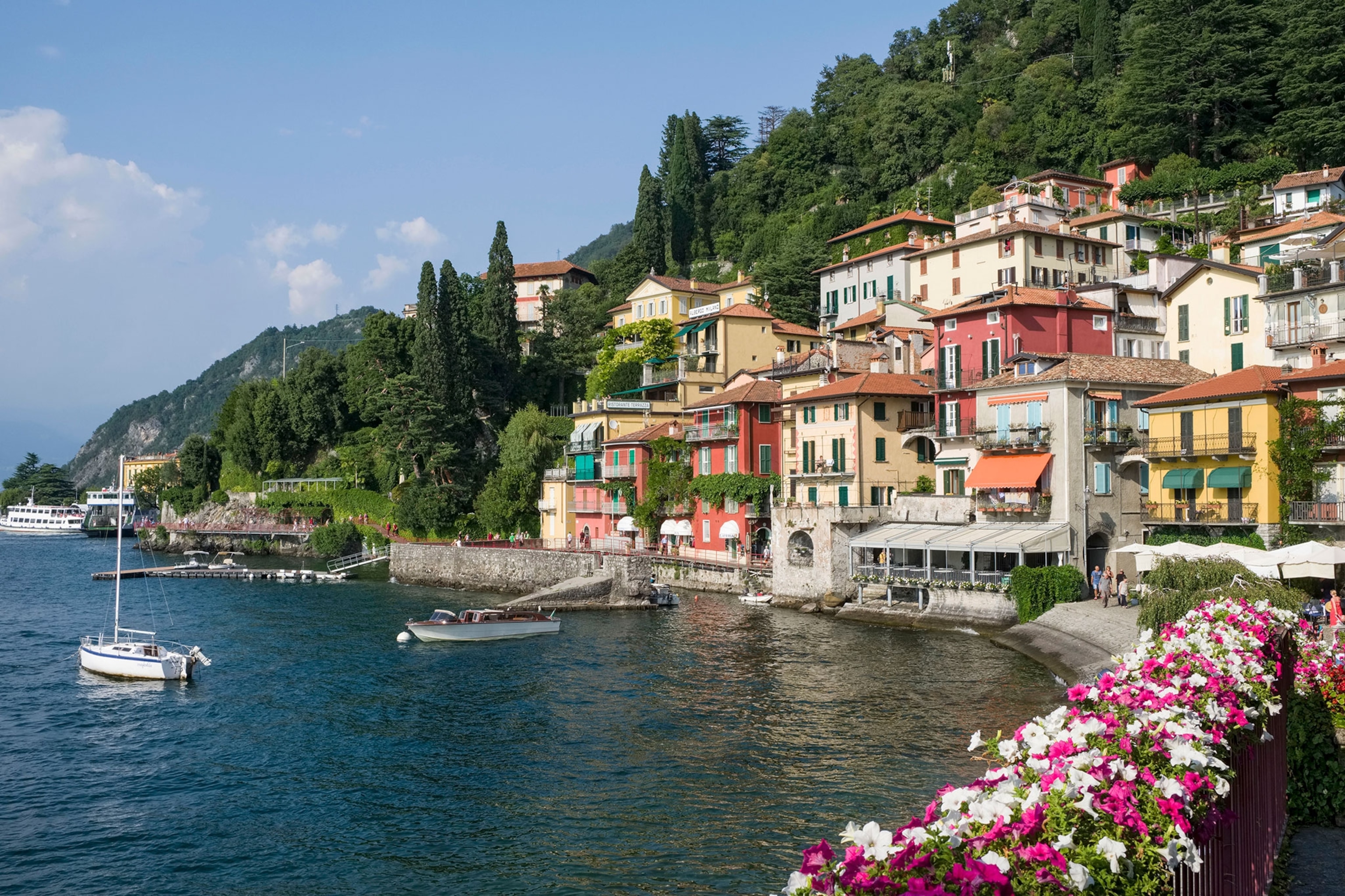 Colorful buildings sit lakeside in front of a mountain filled with green trees. To the left of the houses is a blue lake with boats floating.