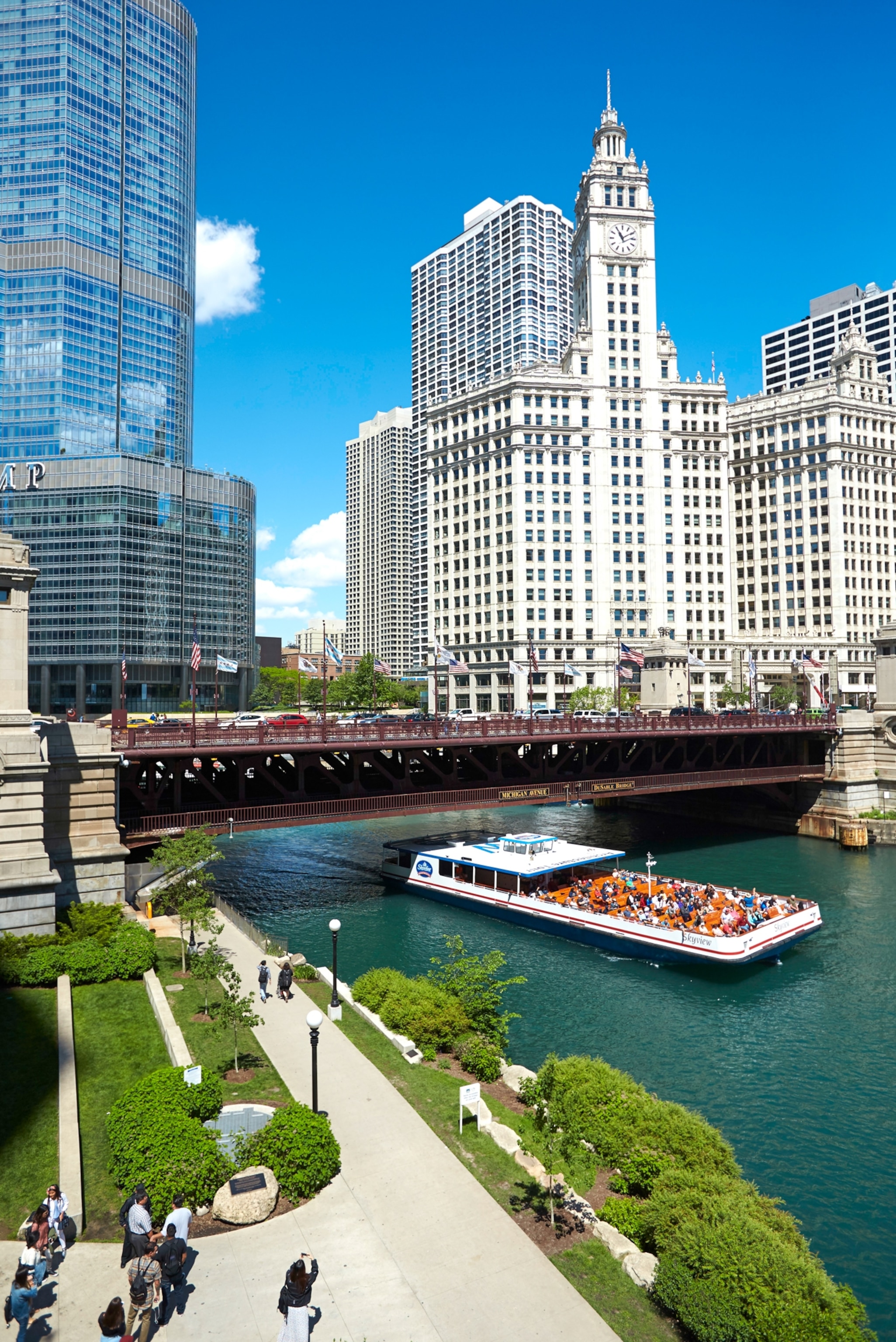 A red ferry next to a boardwalk on a blue river, the cities buildings tower over the space filling the frame.
