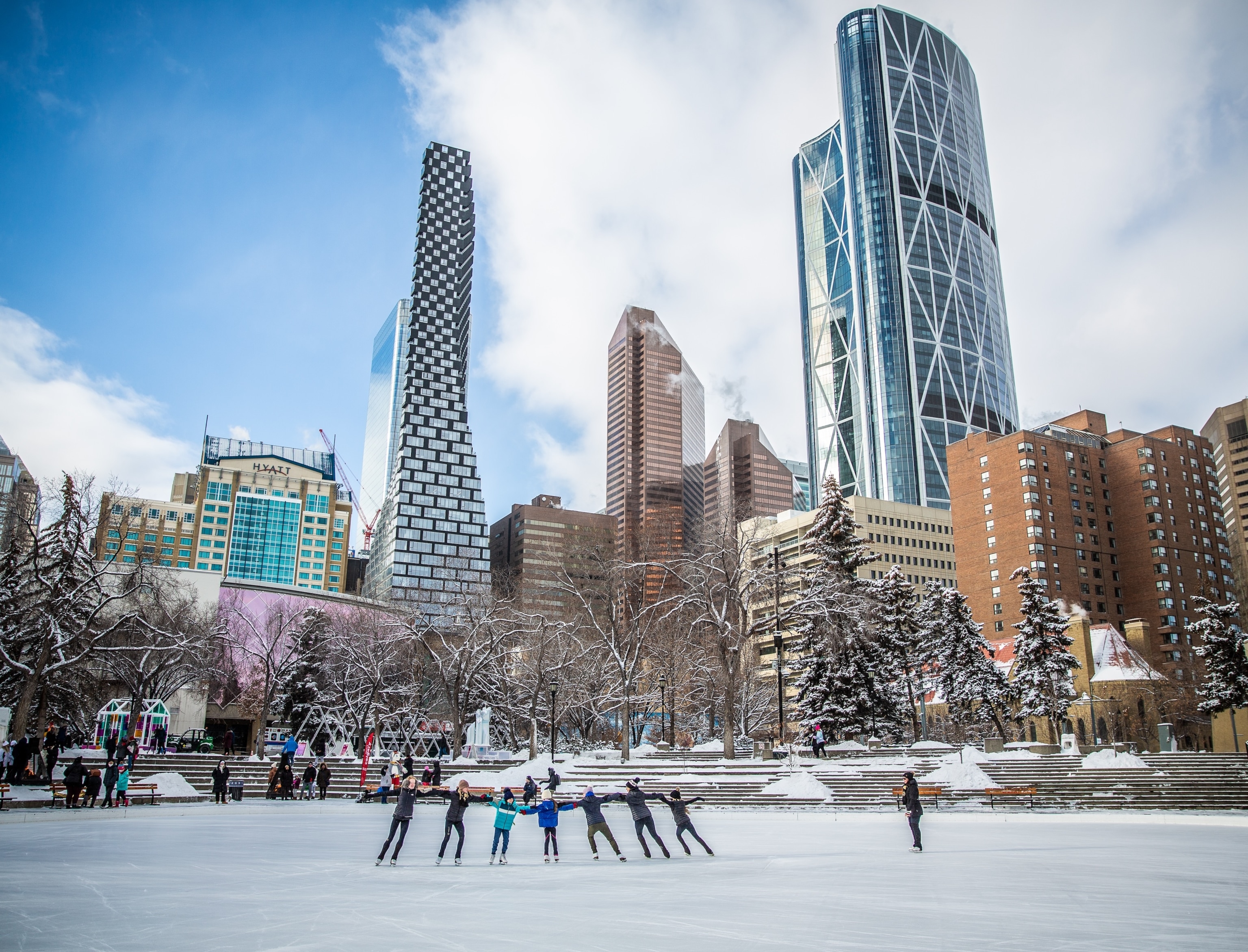Several people ice skate arm in arm in an outdoor ice skating rink in winter surrounded by tall skyscrapers.